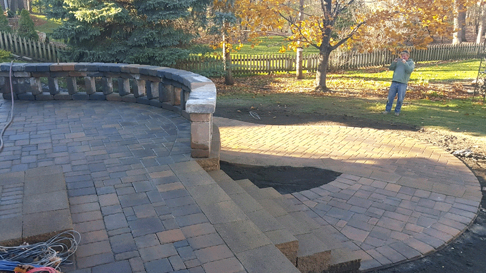 A person stands in a yard next to a newly constructed stone patio featuring a curved retaining wall and stone steps.