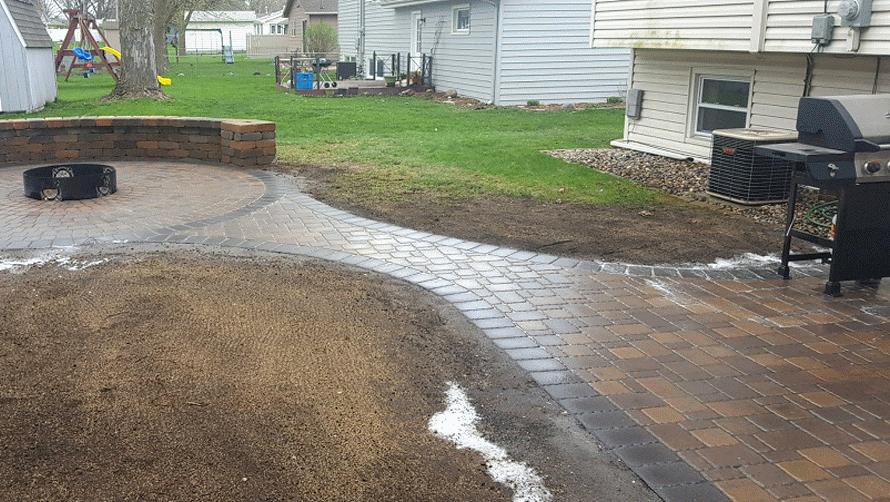 A backyard featuring a newly installed paver patio and walkway connecting a circular fire pit area to a house.
