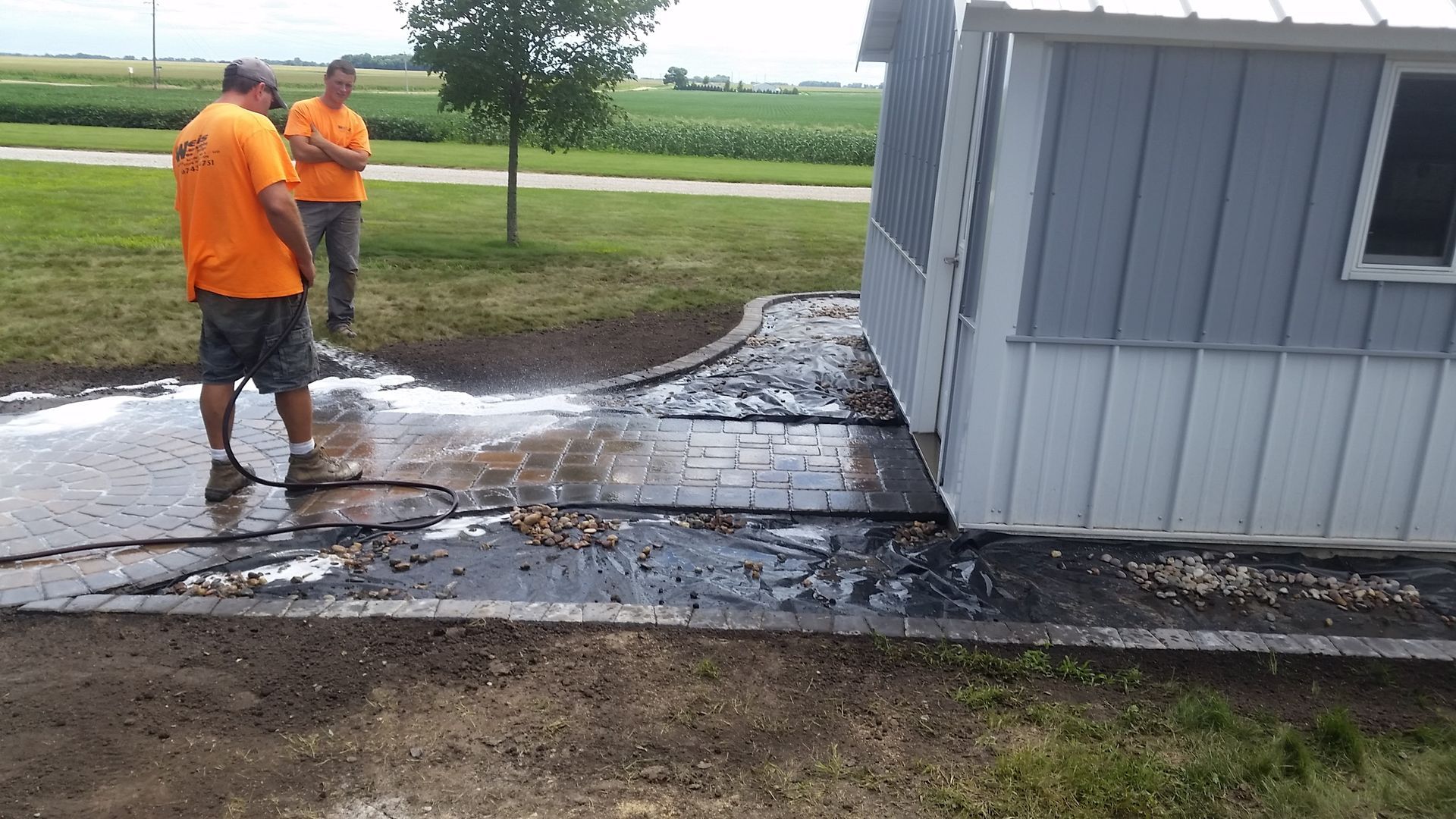 Two workers in bright orange shirts stand on a patio beside a small grey shed while cleaning a paved walkway.