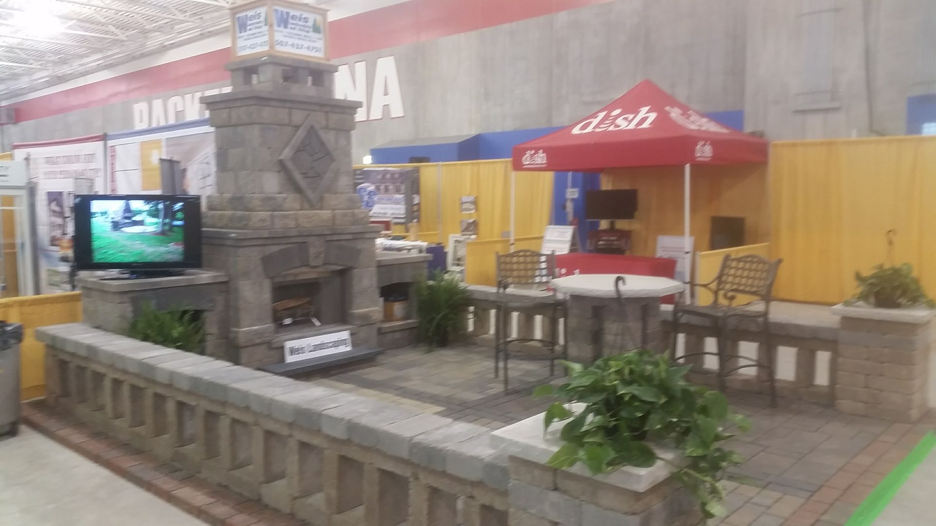 An outdoor living display with a stone fireplace, a small patio, a table with chairs, and a red Dish satellite canopy.