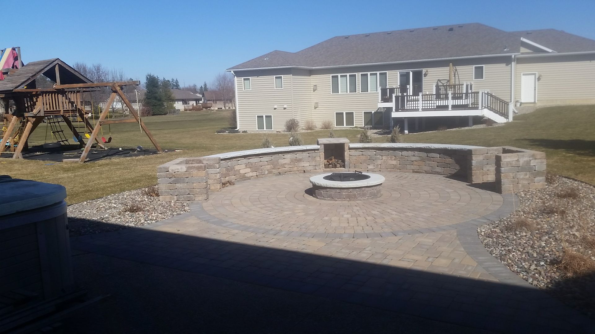 A backyard patio featuring a circular stone fire pit and a curved retaining wall, with a play set and house in the back.
