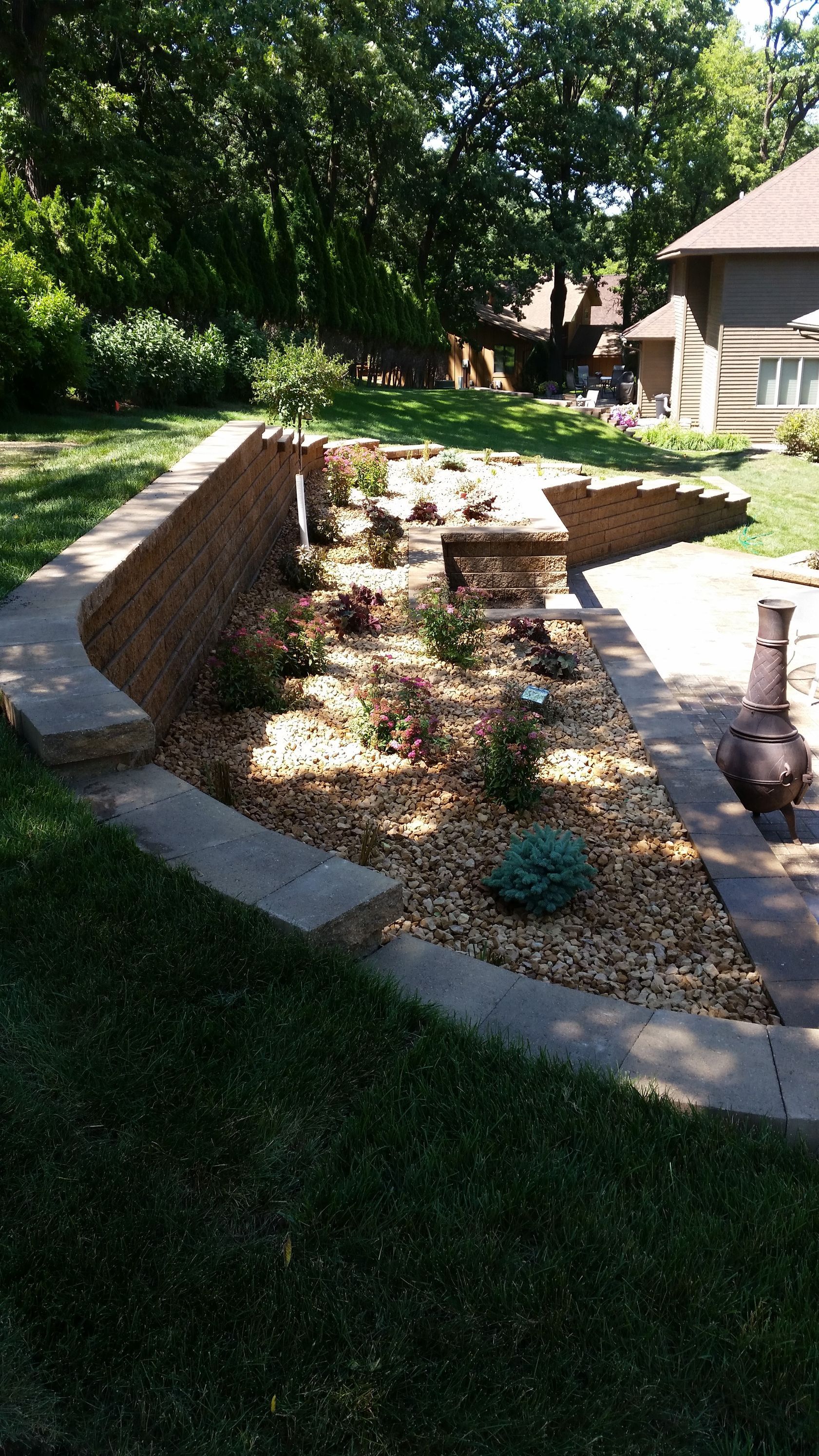 A curved stone retaining wall borders a backyard garden bed filled with light-colored gravel and several small shrubs.