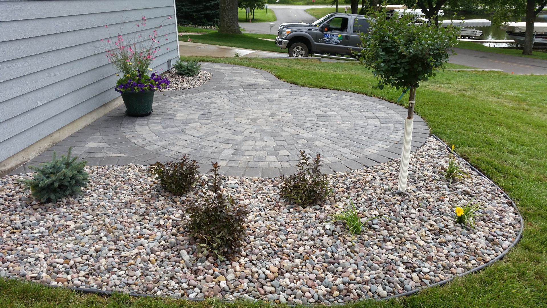 A circular gray paver patio set next to a house wall, surrounded by a gravel garden bed with small shrubs and a tree.