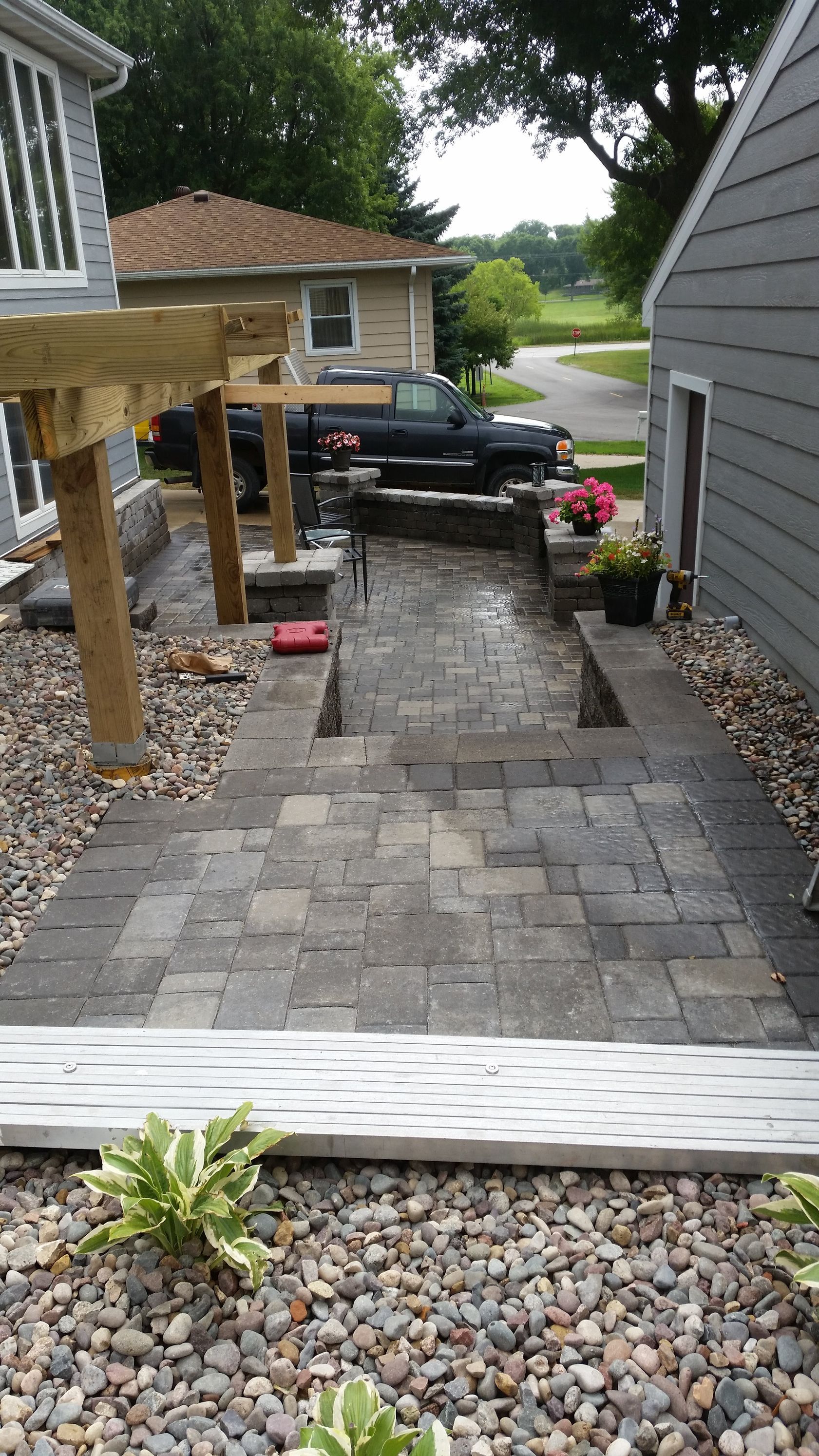 A stone paver walkway leads down from a wooden deck structure toward a backyard with gravel, plants, and a parked truck.