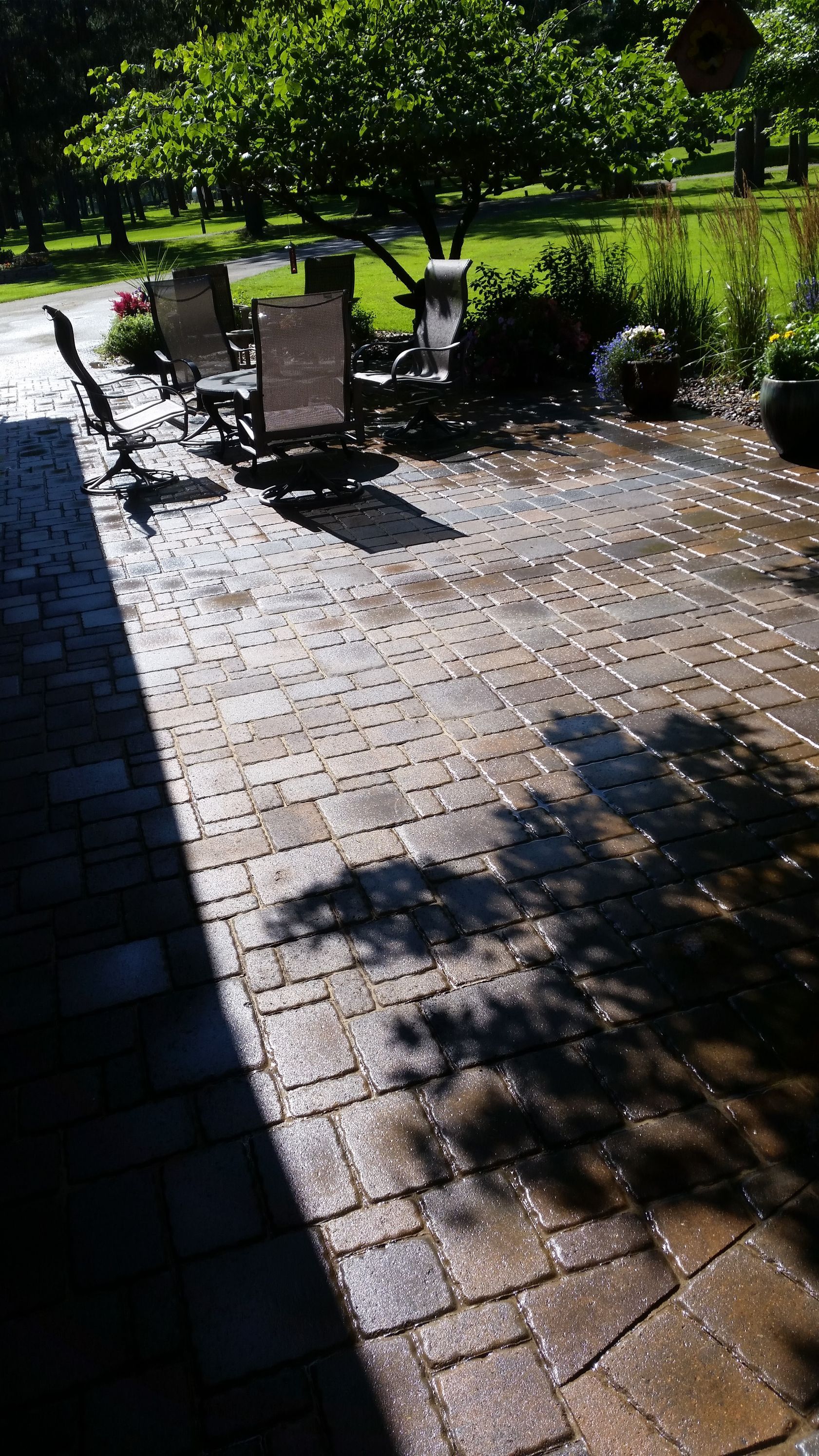 A sunny stone patio with outdoor furniture, shaded by a large tree, leading to a green lawn.