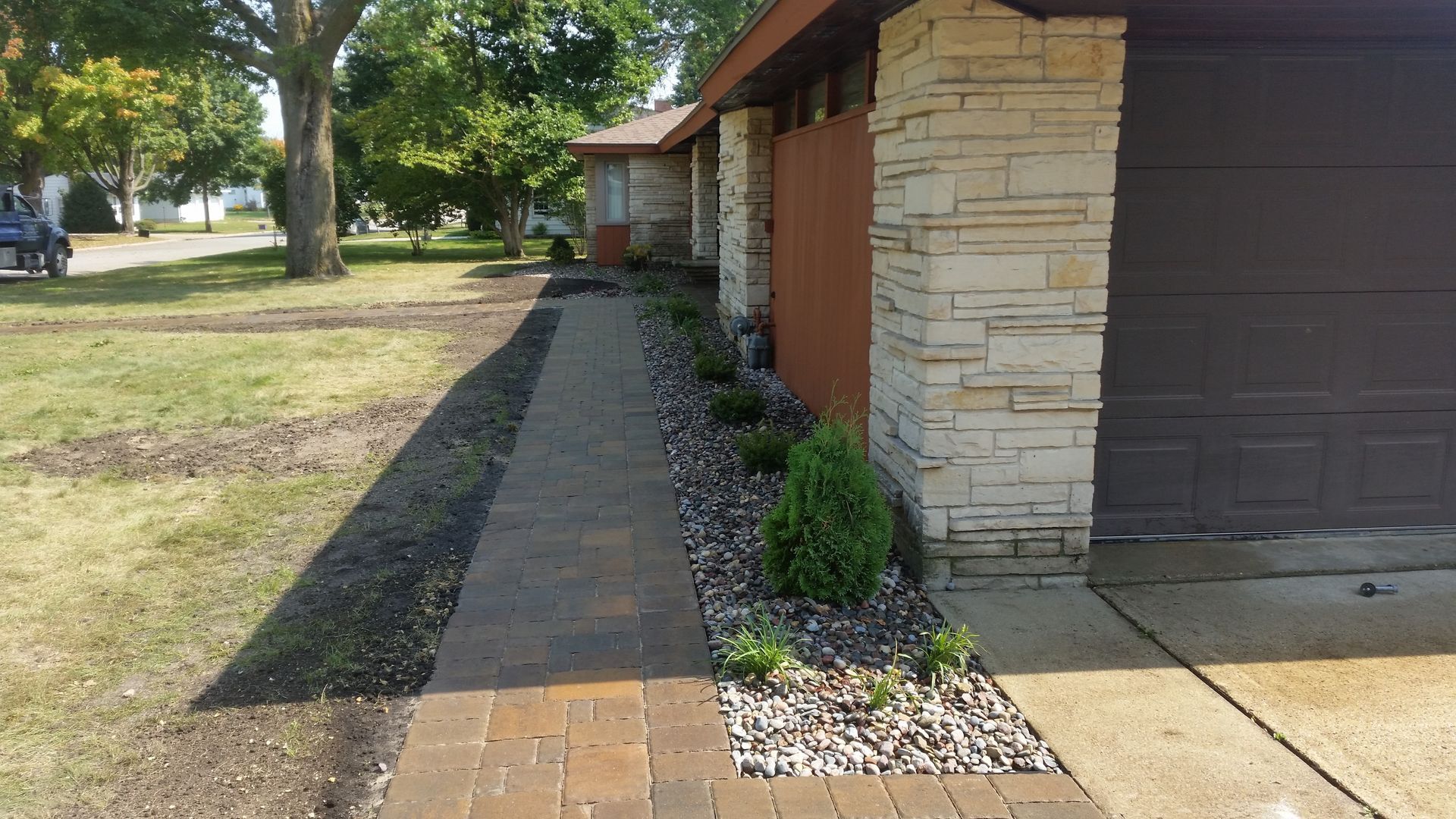 A brown paver walkway leads to a house entrance, flanked by a gravel garden bed with small shrubs and a stone pillar.