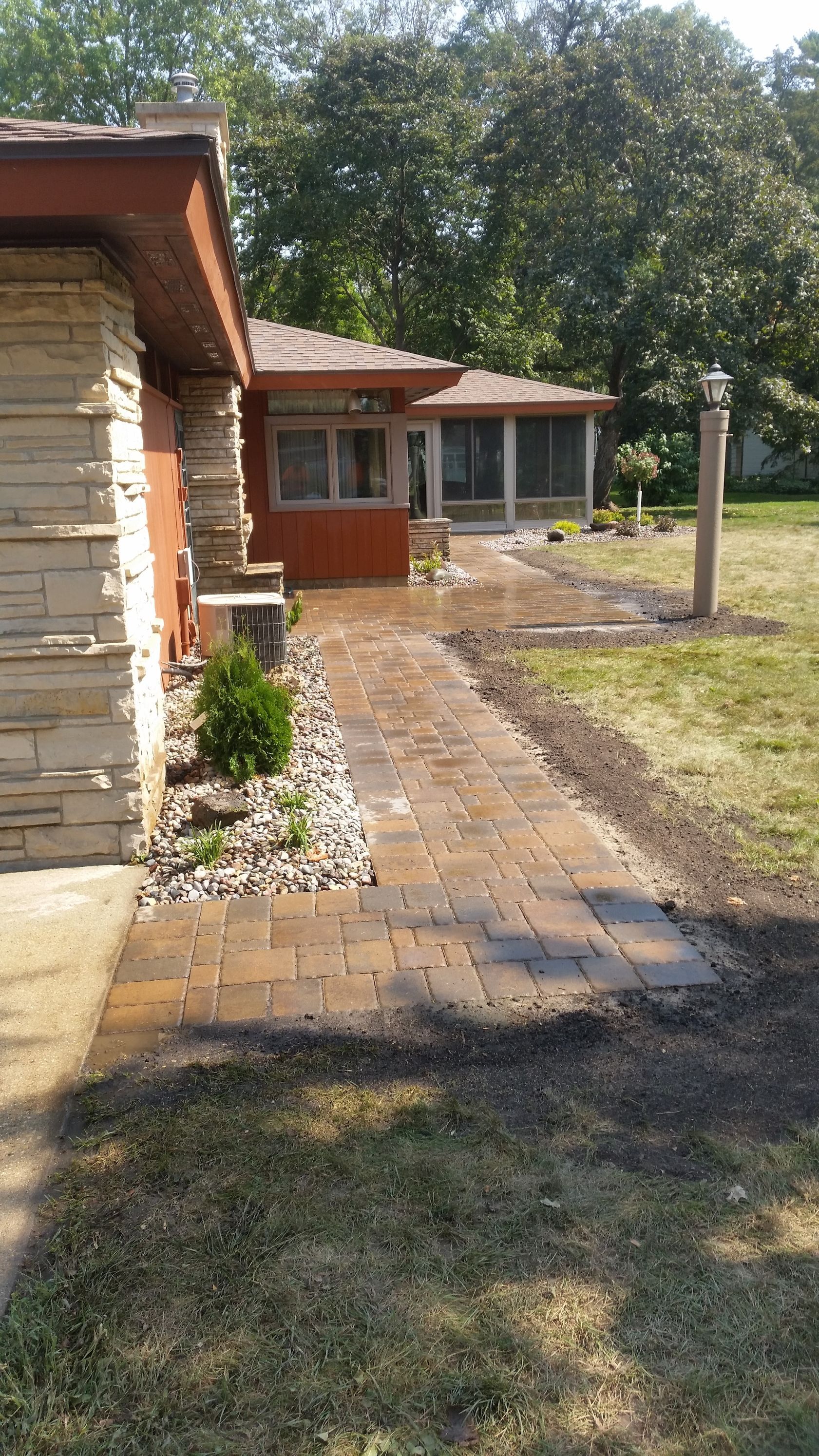 A new tan brick paver walkway leads along the side of a house toward a screened-in porch with brown trim.