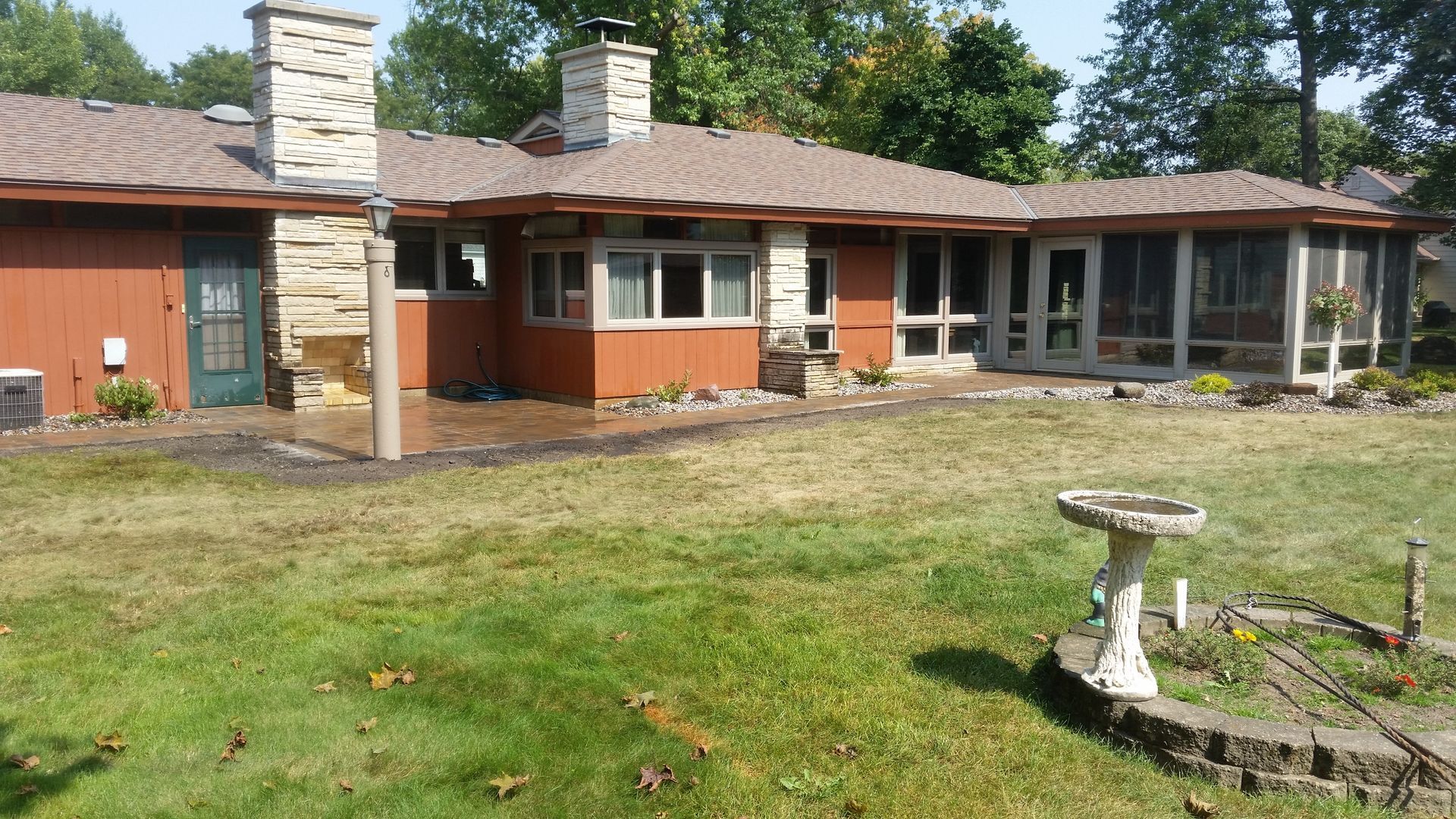 A single-story house with terracotta-colored siding, stone chimneys, and a lawn featuring a birdbath in the foreground.