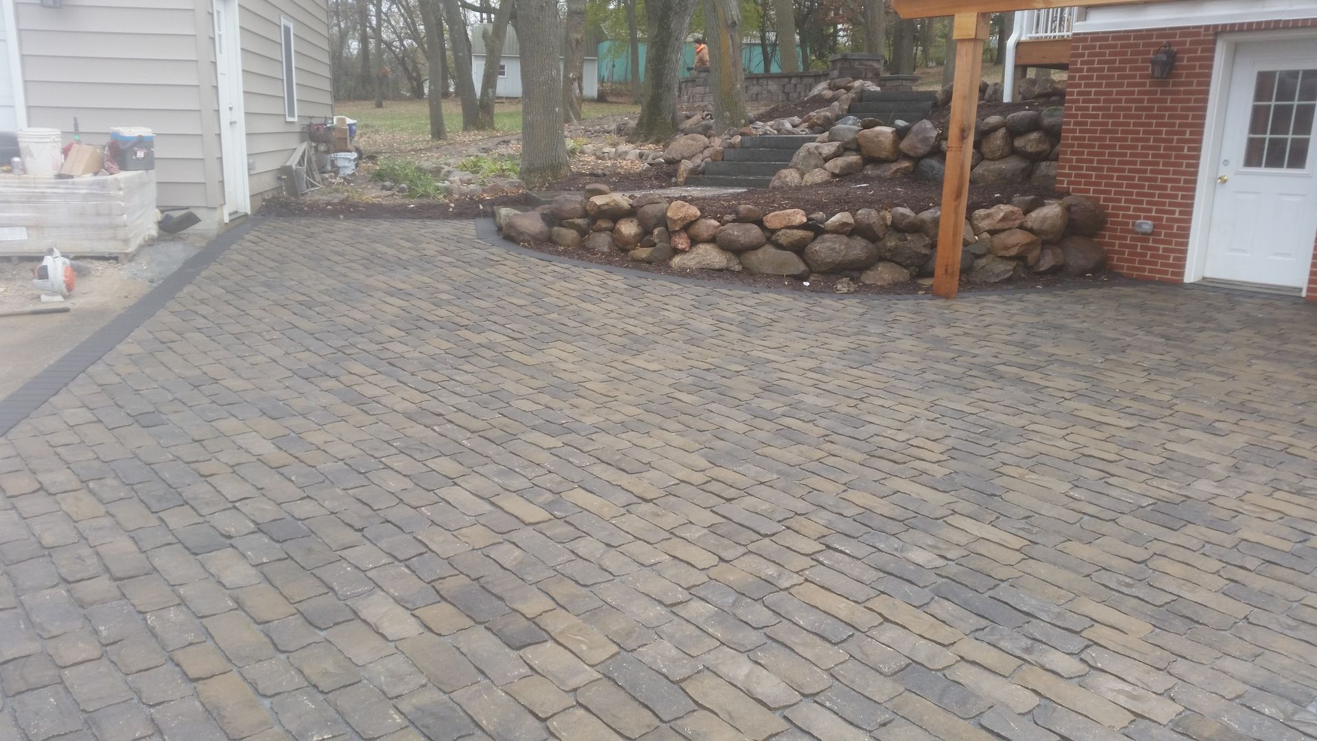 A patio made of multicolored rectangular stone pavers, bordered by a fieldstone retaining wall near a brick house exterior.