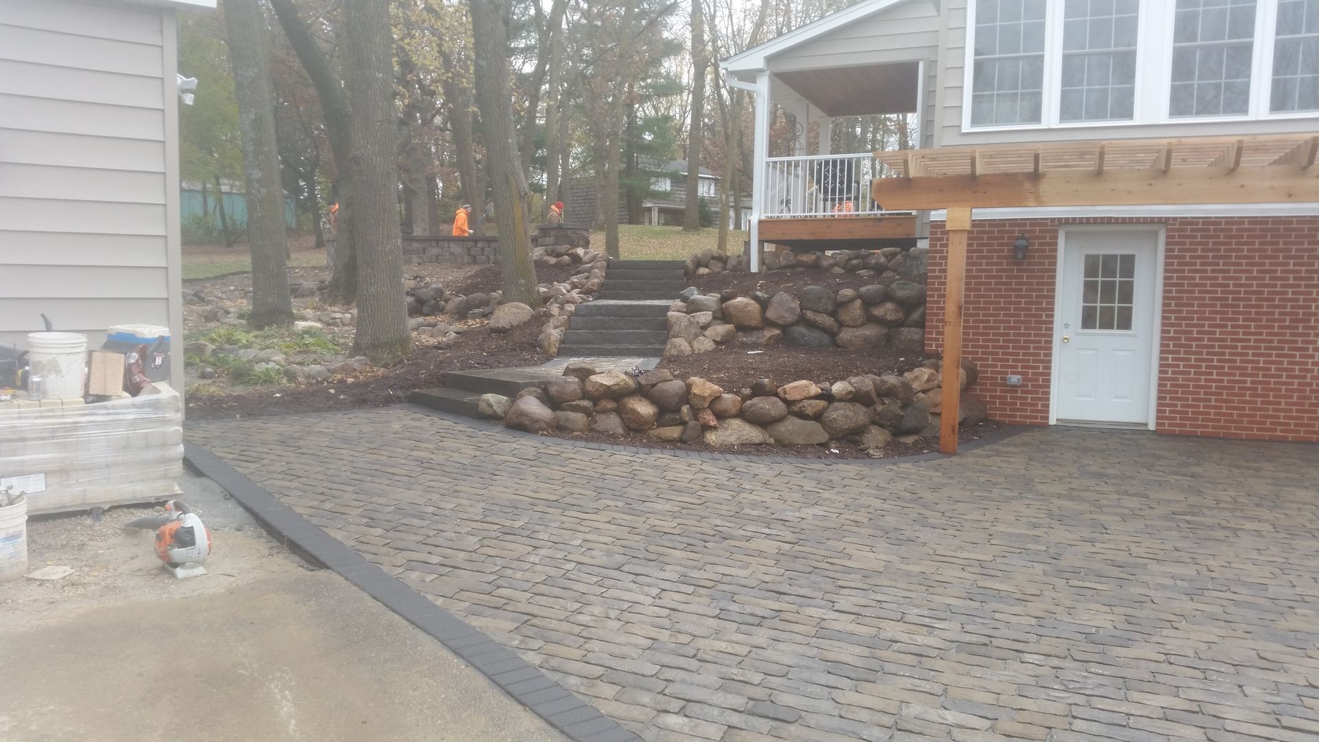 A new brick paver patio sits in front of a house with a brick foundation, rock retaining walls, and outdoor stairs.