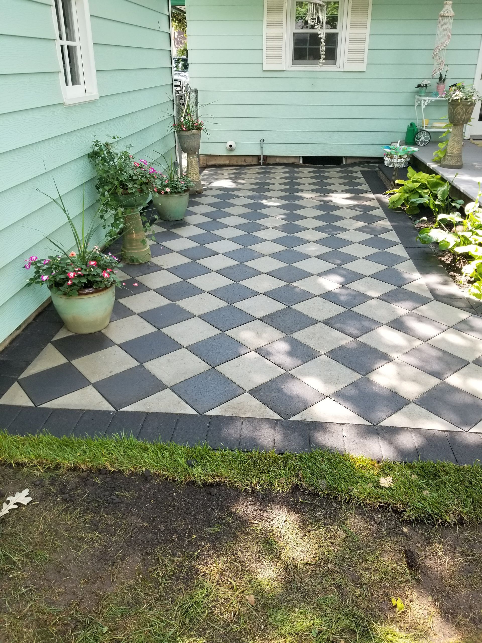 A patio with a gray and white checkered paver design, accented by potted plants against a light green house exterior.