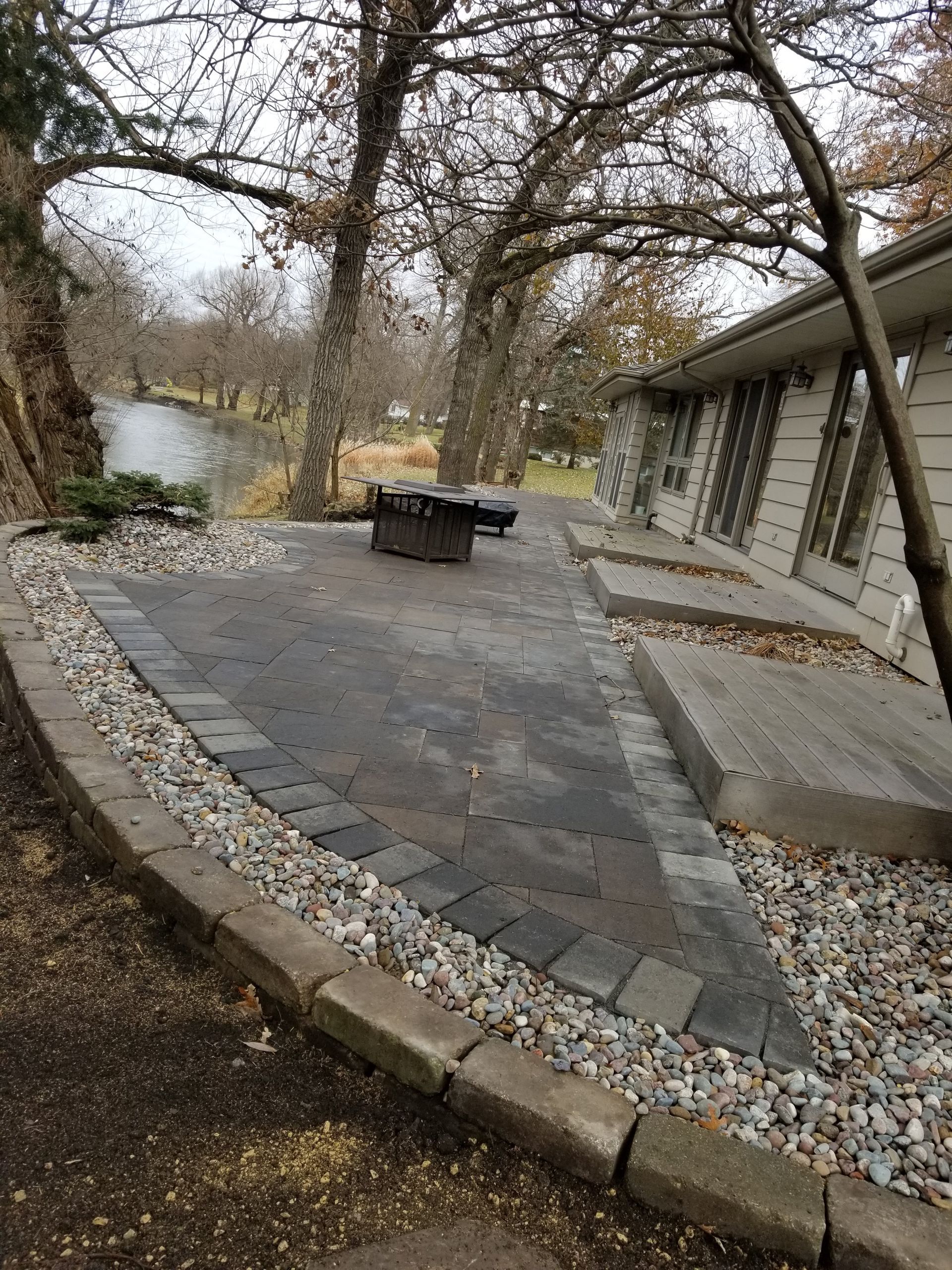 A backyard patio with dark pavers, a fire pit, and light stone borders, next to a house exterior and a river.