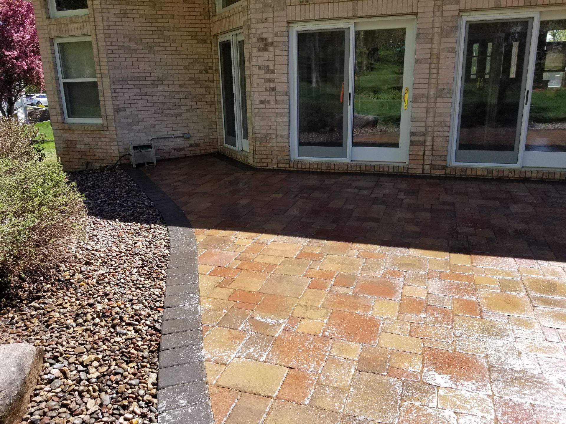 A brick patio with a dark stone border set against a tan brick house with glass sliding doors and a rock garden.