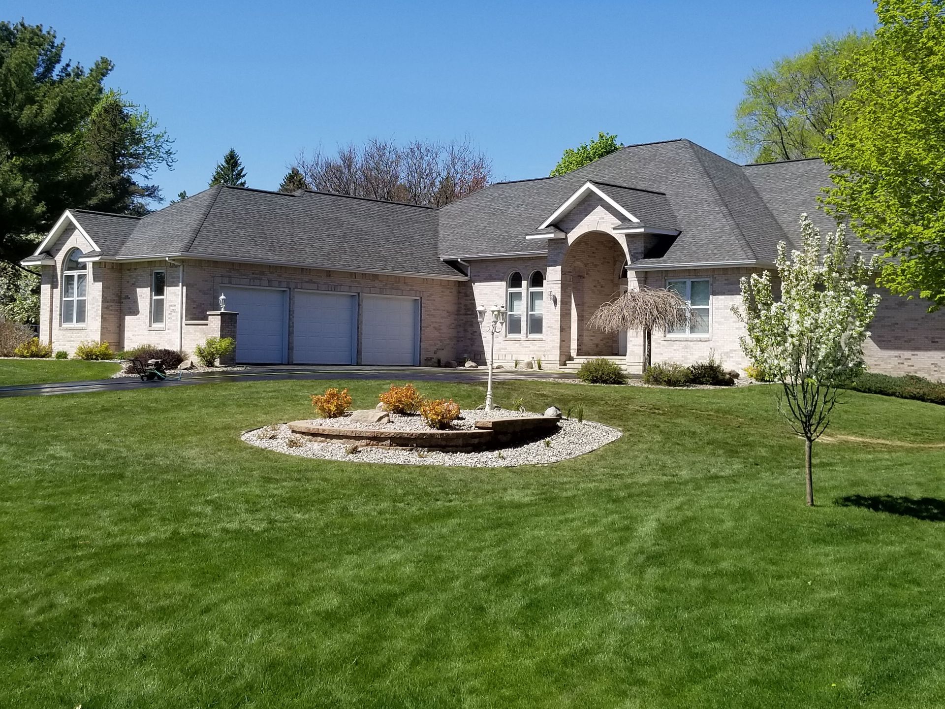 A one-story brick house with a gray roof and three-car garage, featuring a landscaped circular garden in the front lawn.