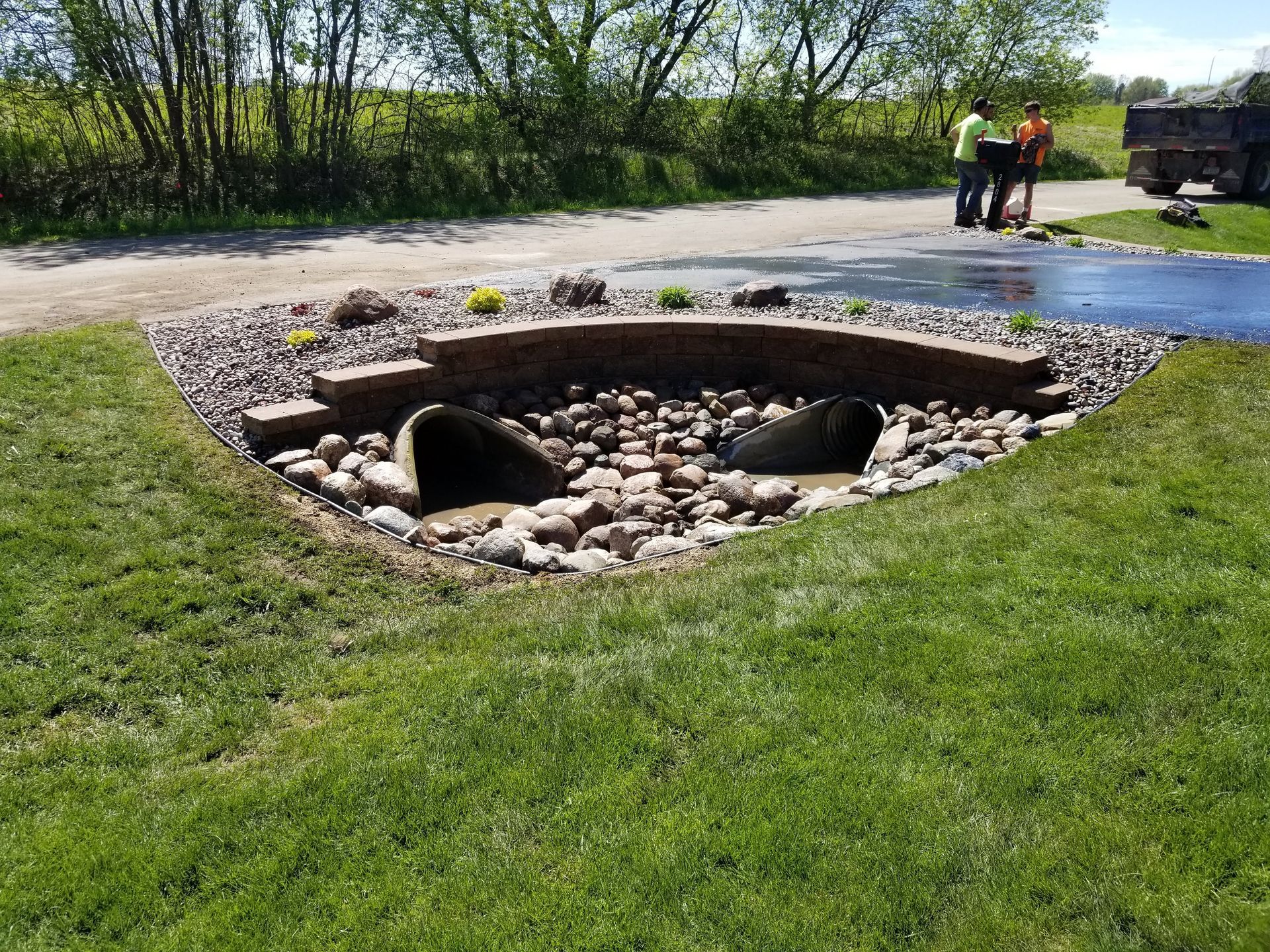 Two workers stand by a drainage area featuring two concrete culverts, a stone retaining wall, and decorative gravel.