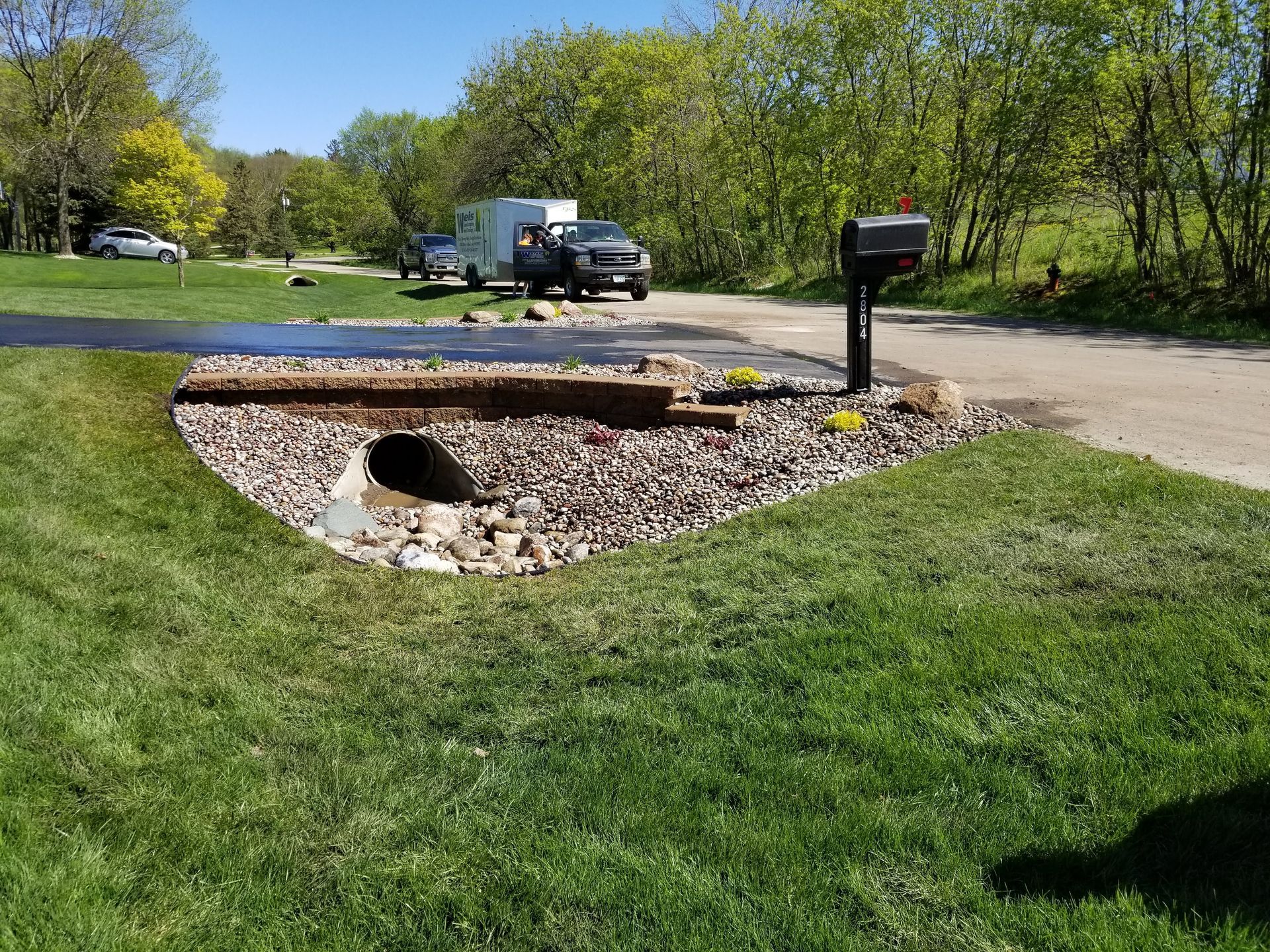 A gravel-filled culvert area with a retaining wall near a mailbox and a driveway in a grassy yard on a sunny day.