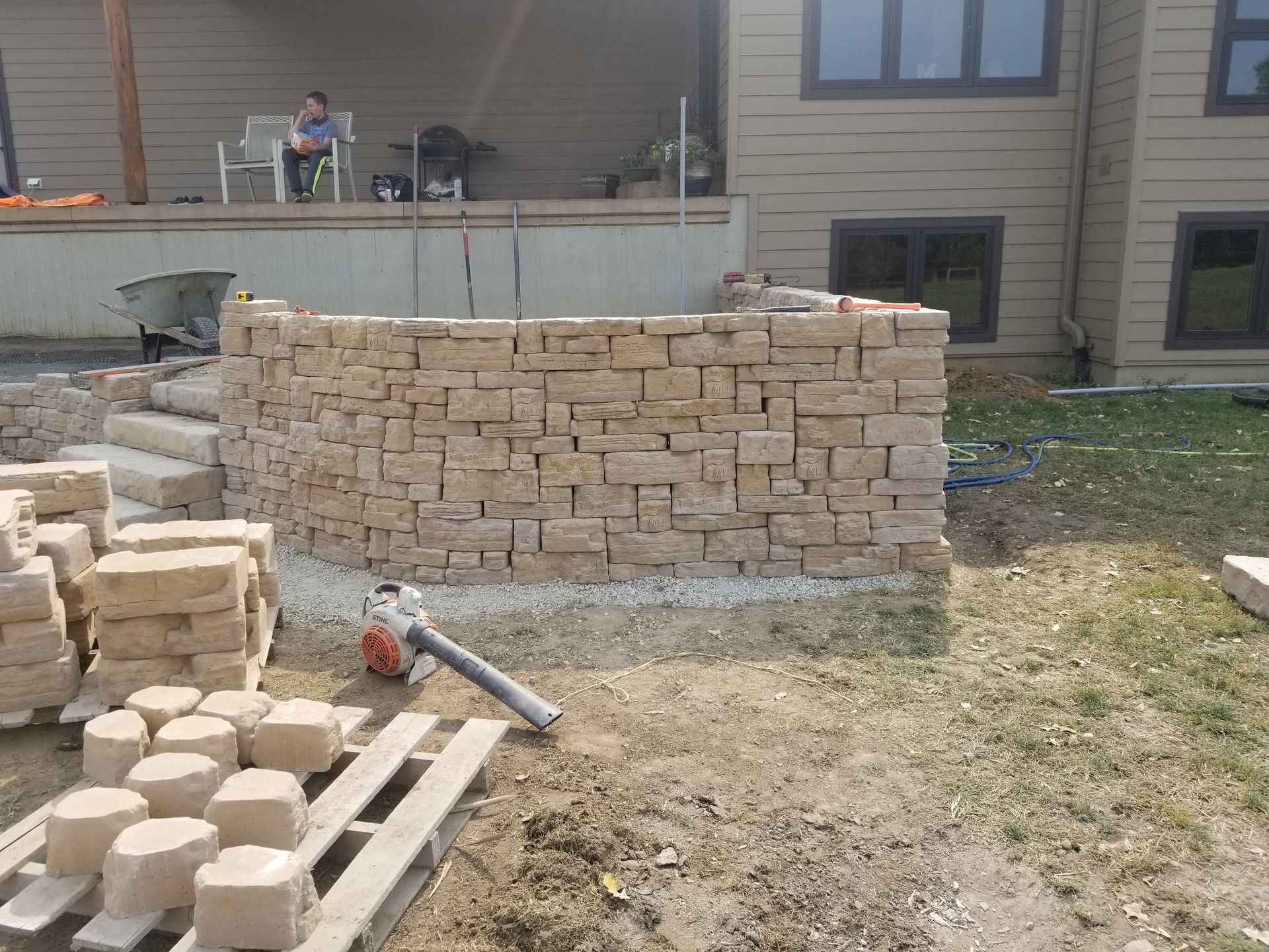 A beige, dry-stacked stone wall under construction in a yard, with stone pallets and a leaf blower in the foreground.