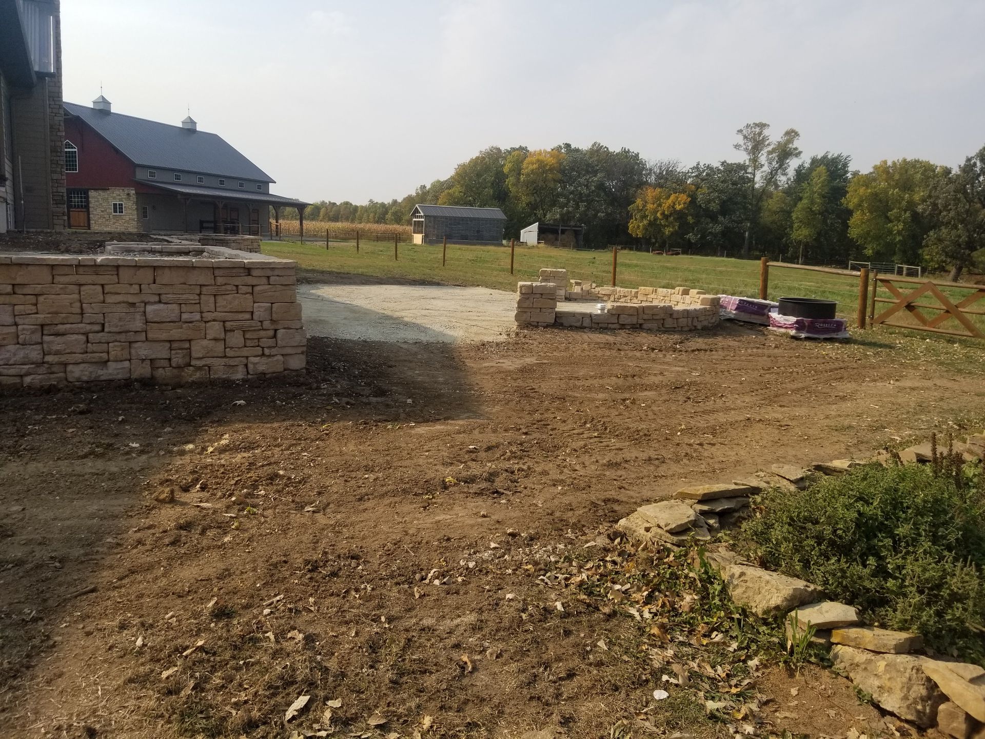 A construction site with a stone retaining wall, an unfinished patio area, and a red barn in the distance.