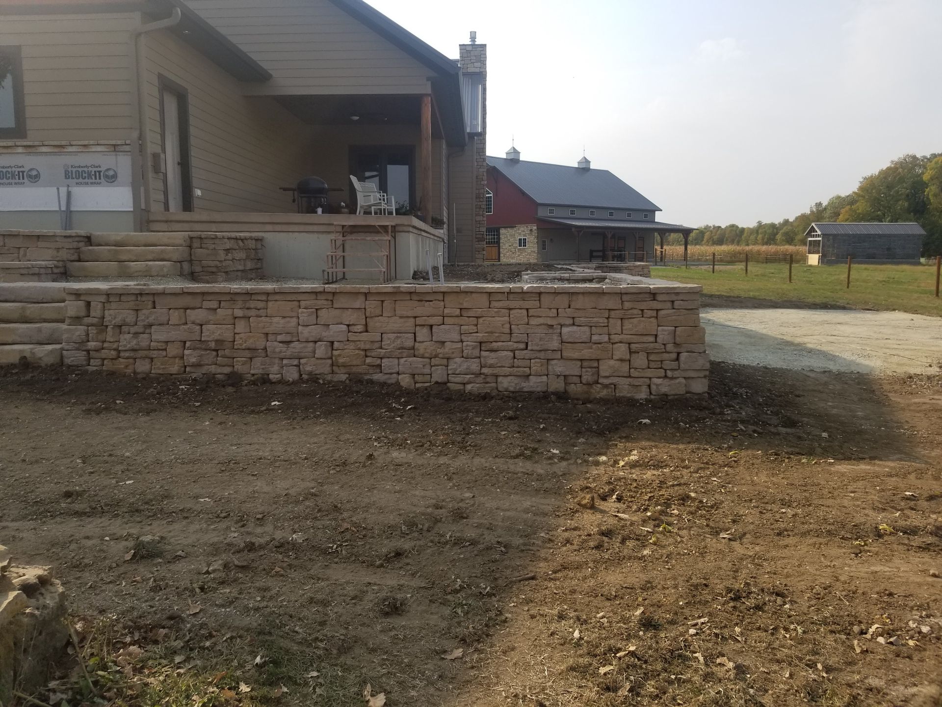 A stone retaining wall sits in front of a house under construction, with a red barn in the distance on a sunny day.