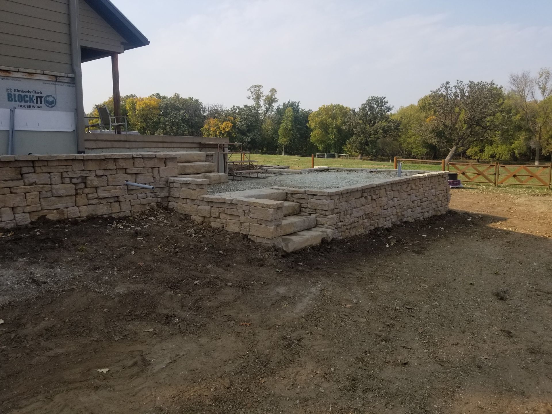 A stone retaining wall and patio base under construction outside a house, with a natural landscape in the background.