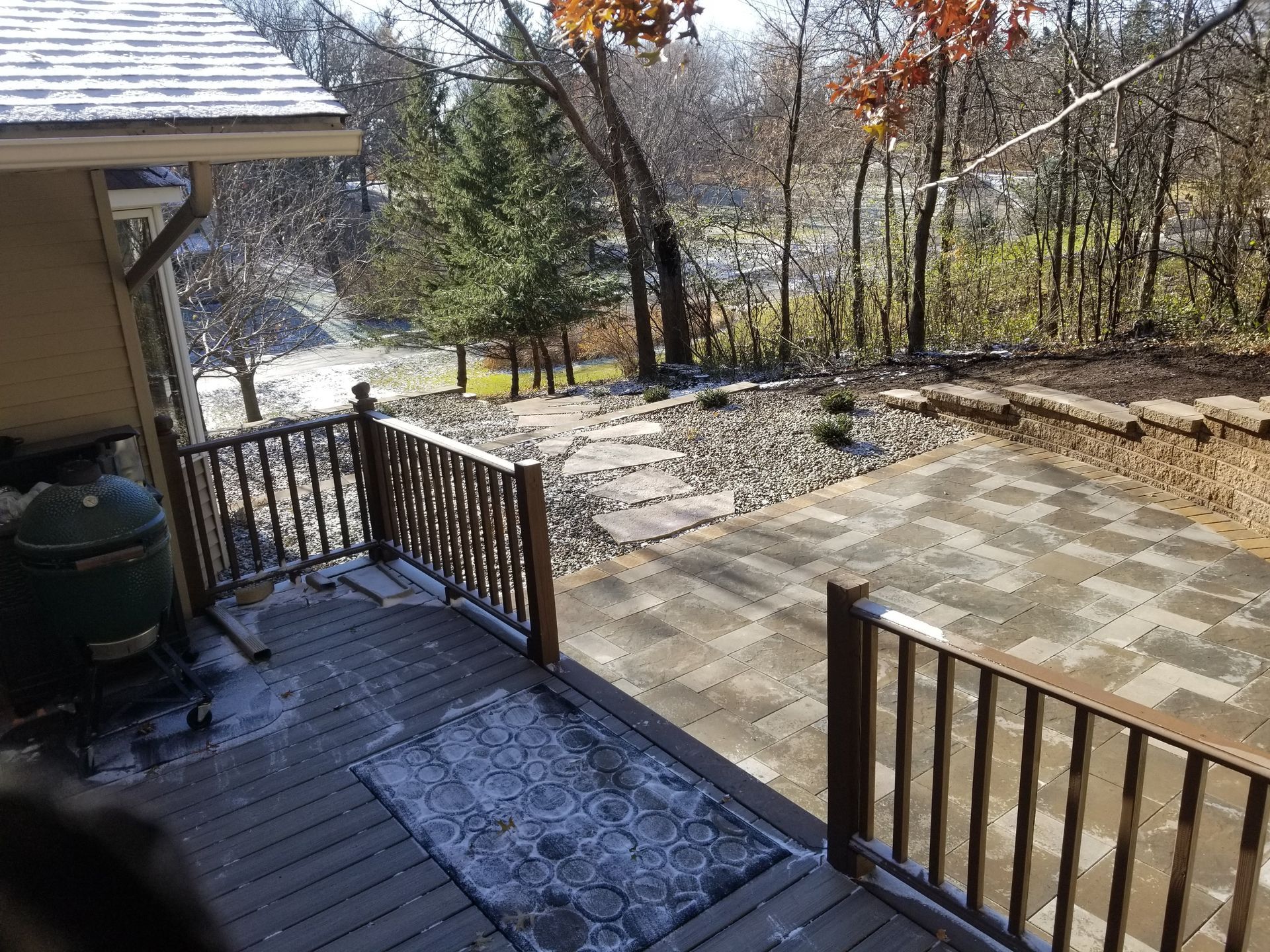 A wooden deck with a grill overlooks a stone patio, a rock garden, and a wooded backyard on a sunny day.