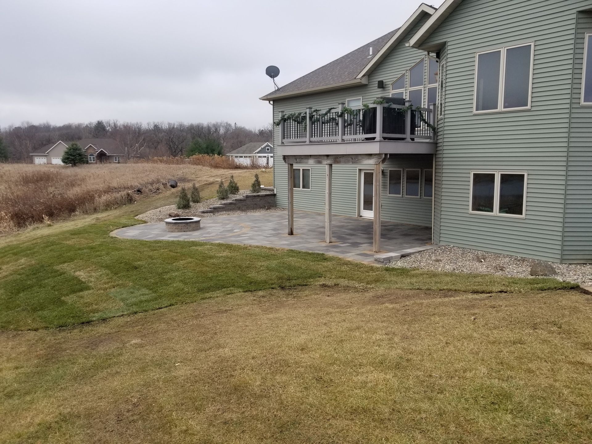 A green two-story house with a wooden deck above a stone patio in a grassy backyard under an overcast sky.