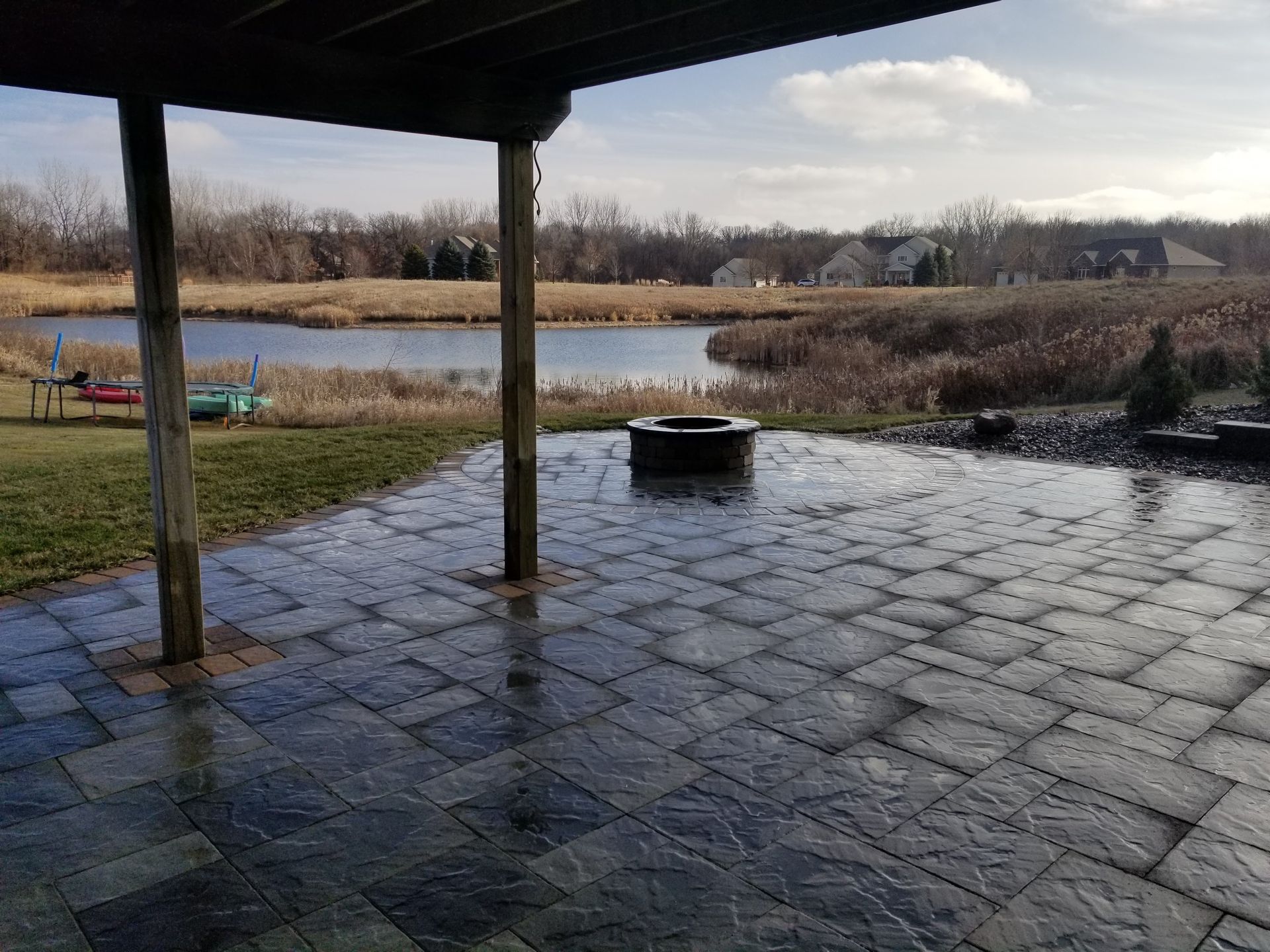 A stone patio with a fire pit overlooking a pond and landscape under a partially cloudy sky.