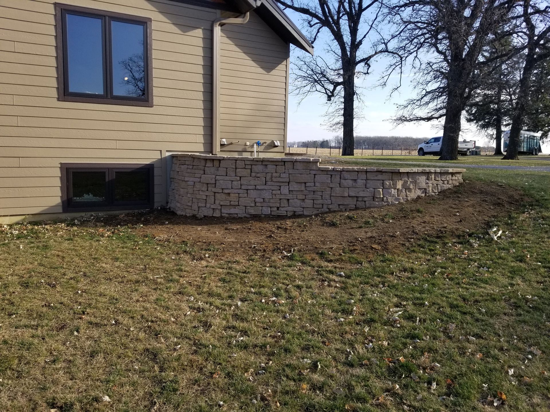 Tan siding house with a basement window and a newly constructed light-colored stone retaining wall in a grassy yard.