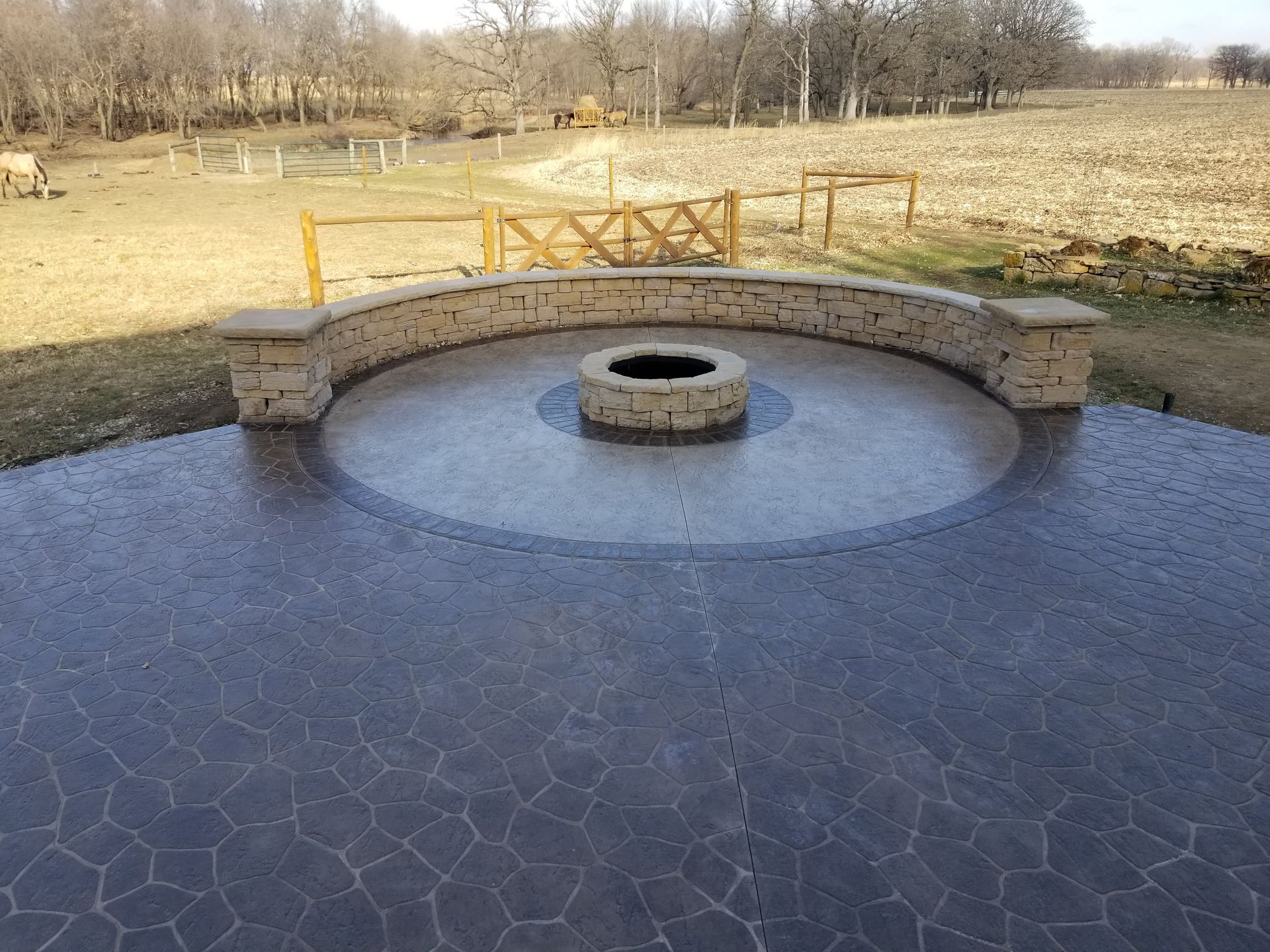 A stone fire pit sits in the center of a circular stamped concrete patio with a curved stone seating wall in a rural yard.