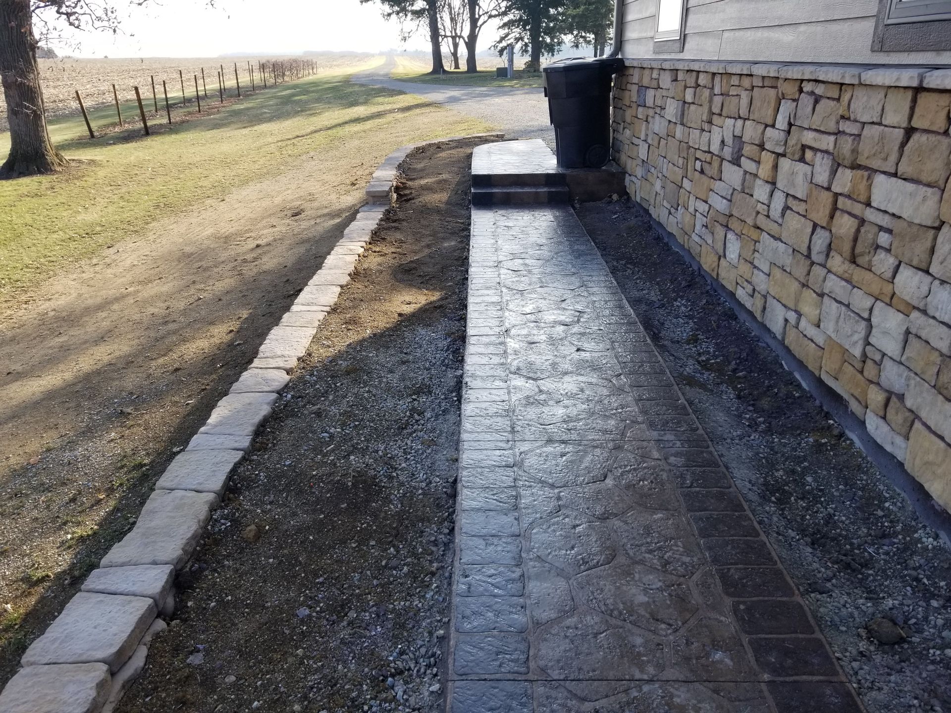 A newly poured, stamped concrete walkway runs along a stone-clad house exterior next to a low, landscaped stone border.