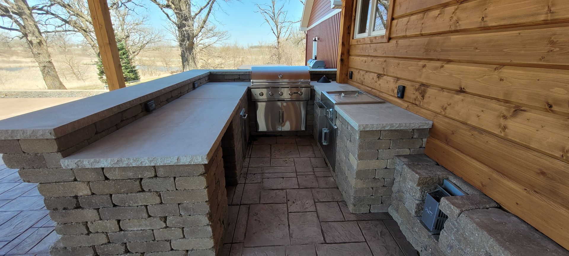 An outdoor kitchen built from stone pavers sits on a patio next to a log cabin, featuring a grill and bar seating area.
