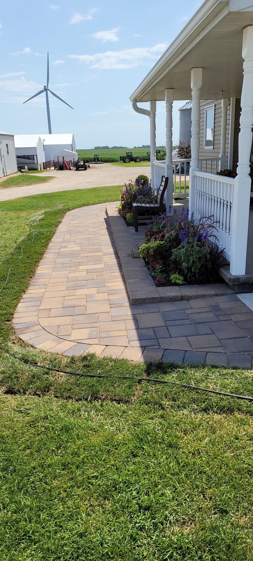 A stone walkway leads to a covered porch with a bench and garden, set on a rural property with a wind turbine in the back.