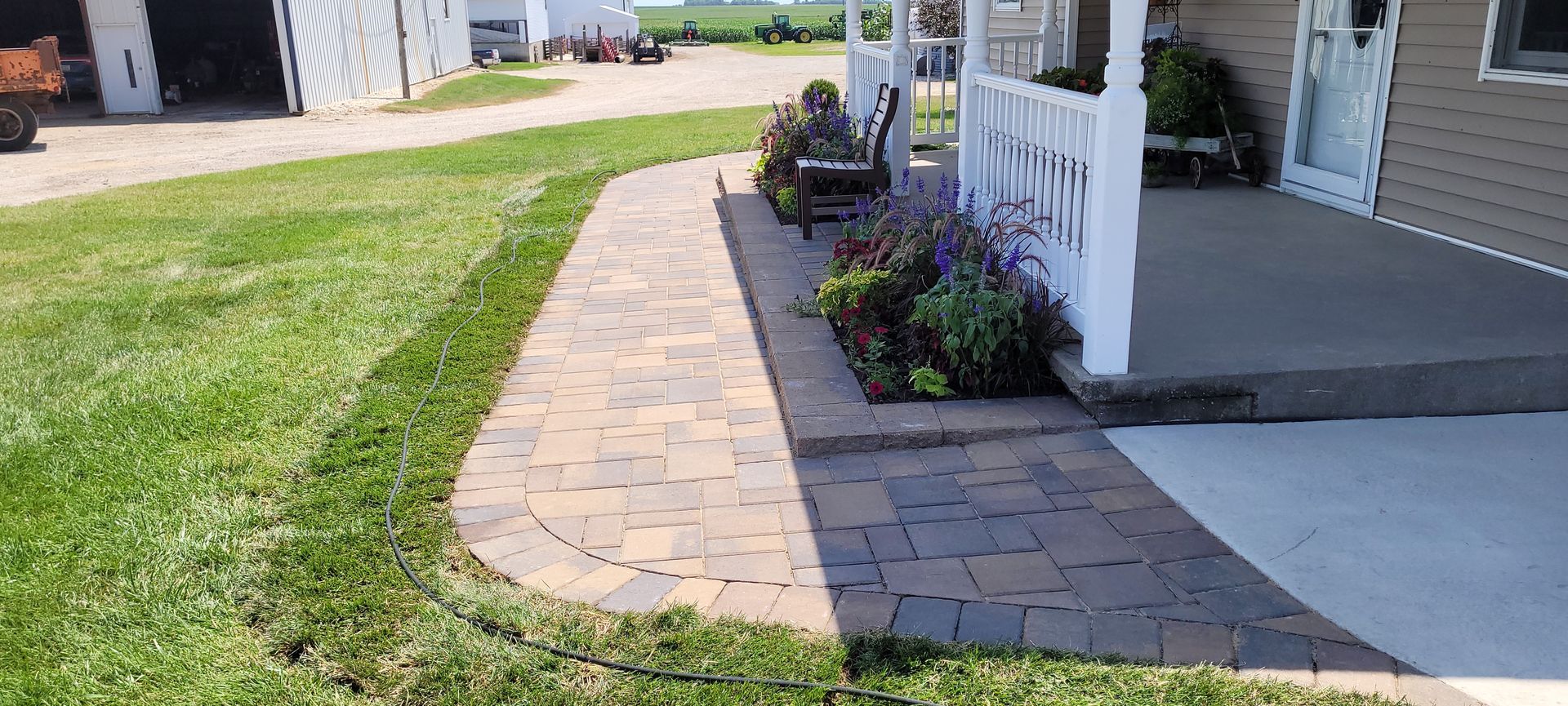 A stone paver walkway curves along the edge of a house porch, featuring a small garden bed and outdoor seating.