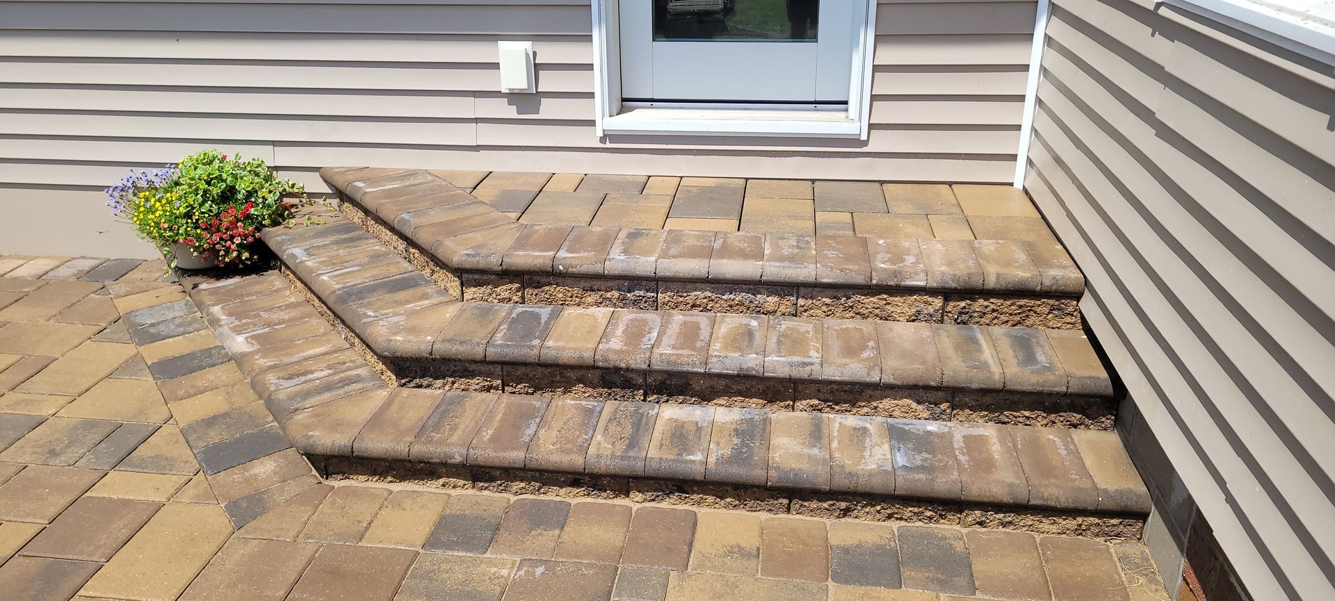 Three stone steps leading up to a doorway, featuring tan, brown, and grey pavers with a potted plant on the left.