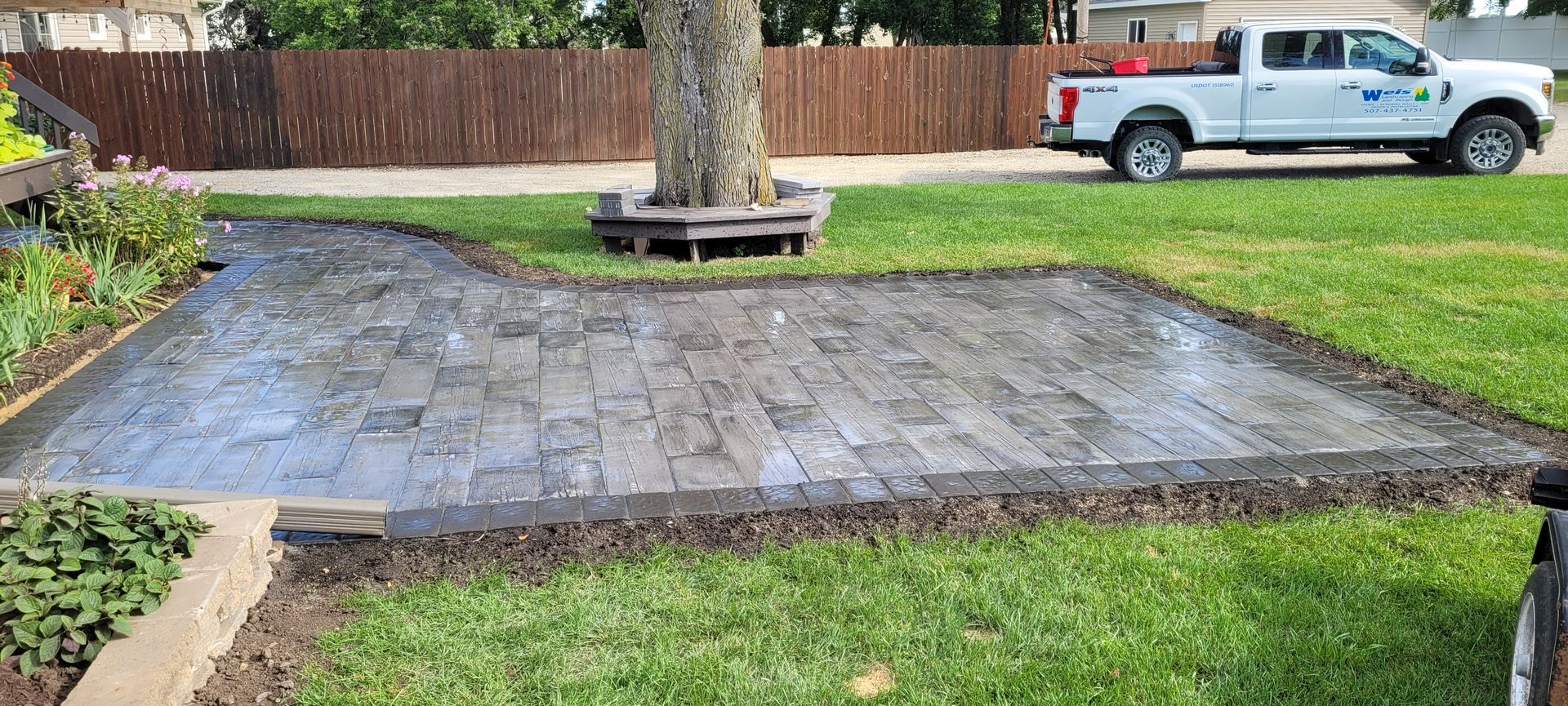 A freshly installed rectangular stone patio in a grassy yard, with a tree in the center and a white truck in the background.