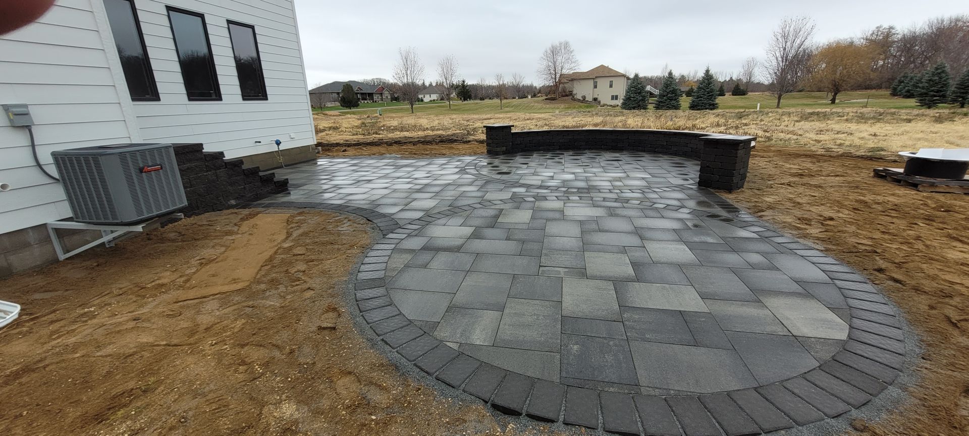 A newly installed gray stone patio with a curved retaining wall bordering a backyard under a cloudy sky.