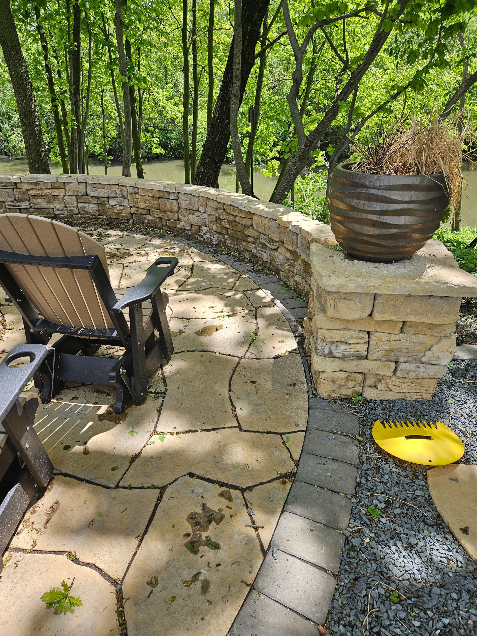 A patio chair sits on a stone-paved terrace overlooking a wooded area and a river, with a planter on a stone pillar.