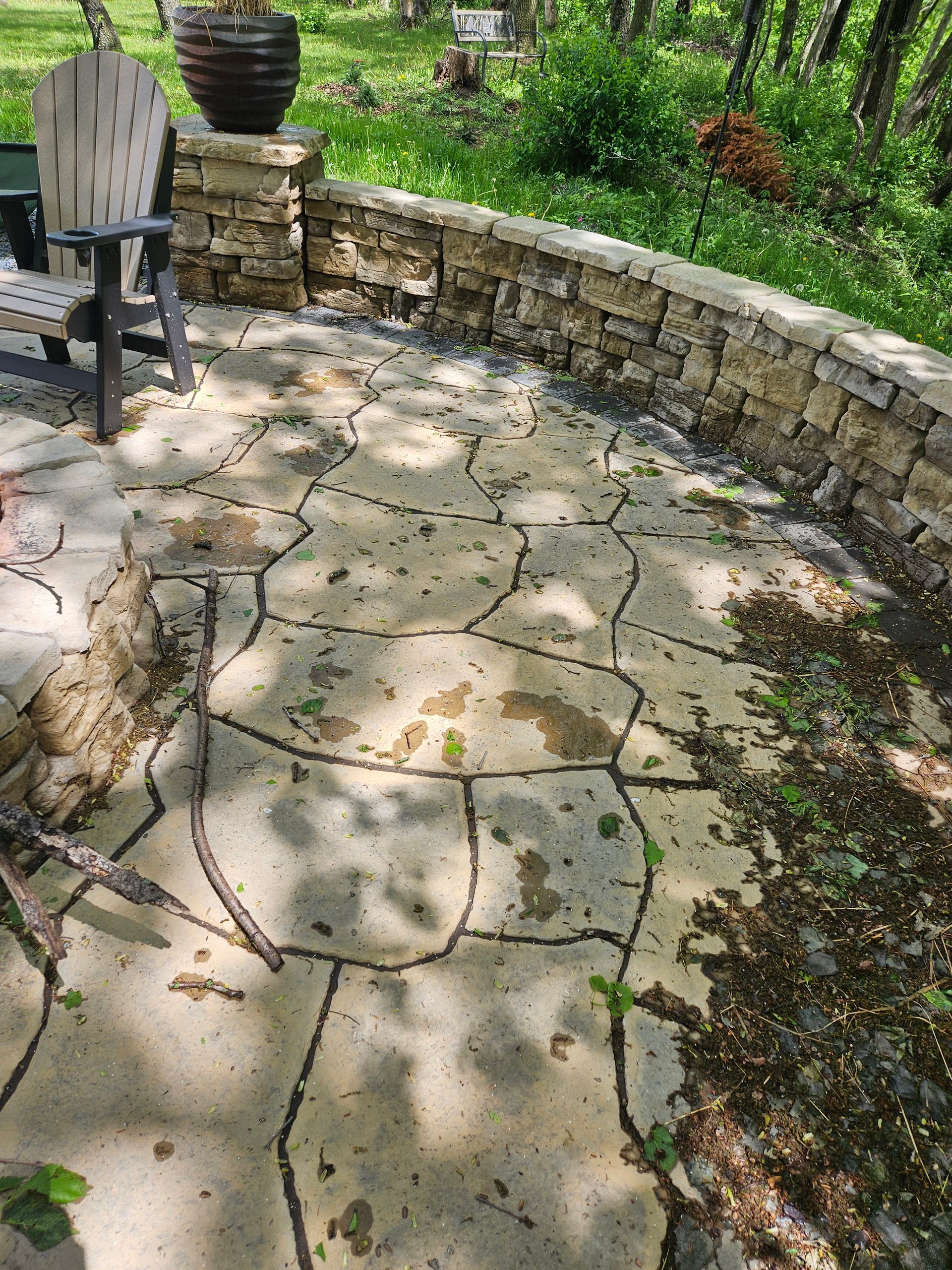 A flagstone patio featuring a stone retaining wall, outdoor chair, and a planter in a wooded backyard.