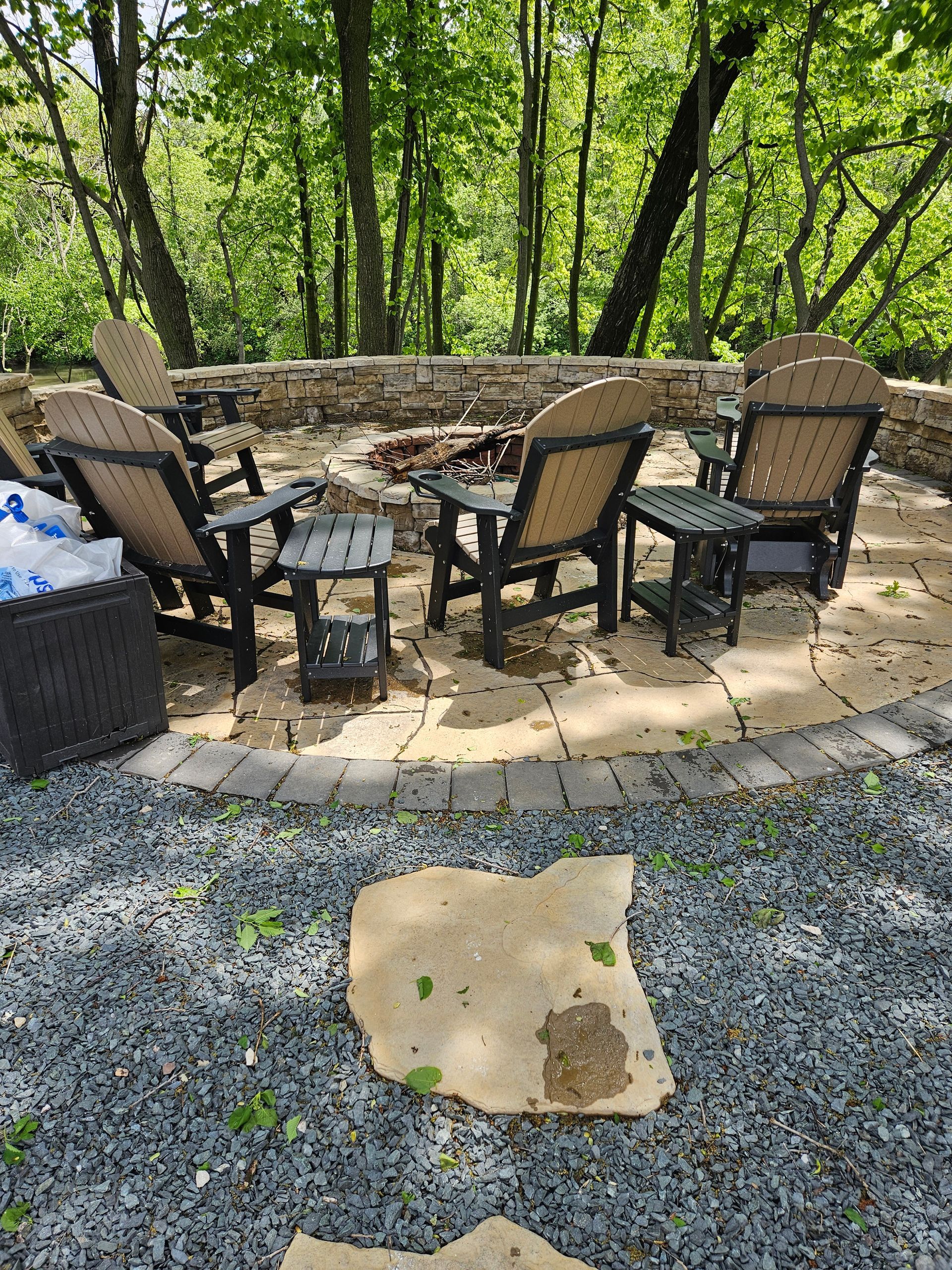 A cozy stone fire pit area in a wooded backyard, featuring four outdoor chairs and side tables on a paved patio.