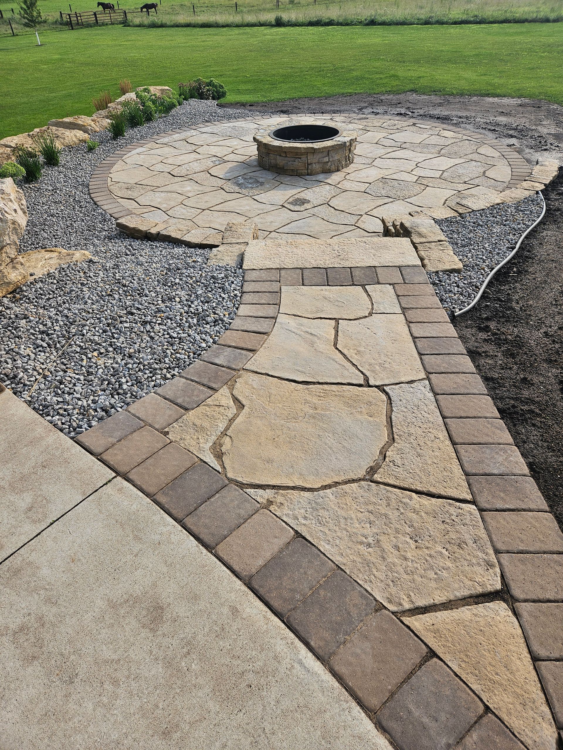 A stone walkway leads to a circular flagstone patio with a stone fire pit, surrounded by gray gravel and a grassy lawn.