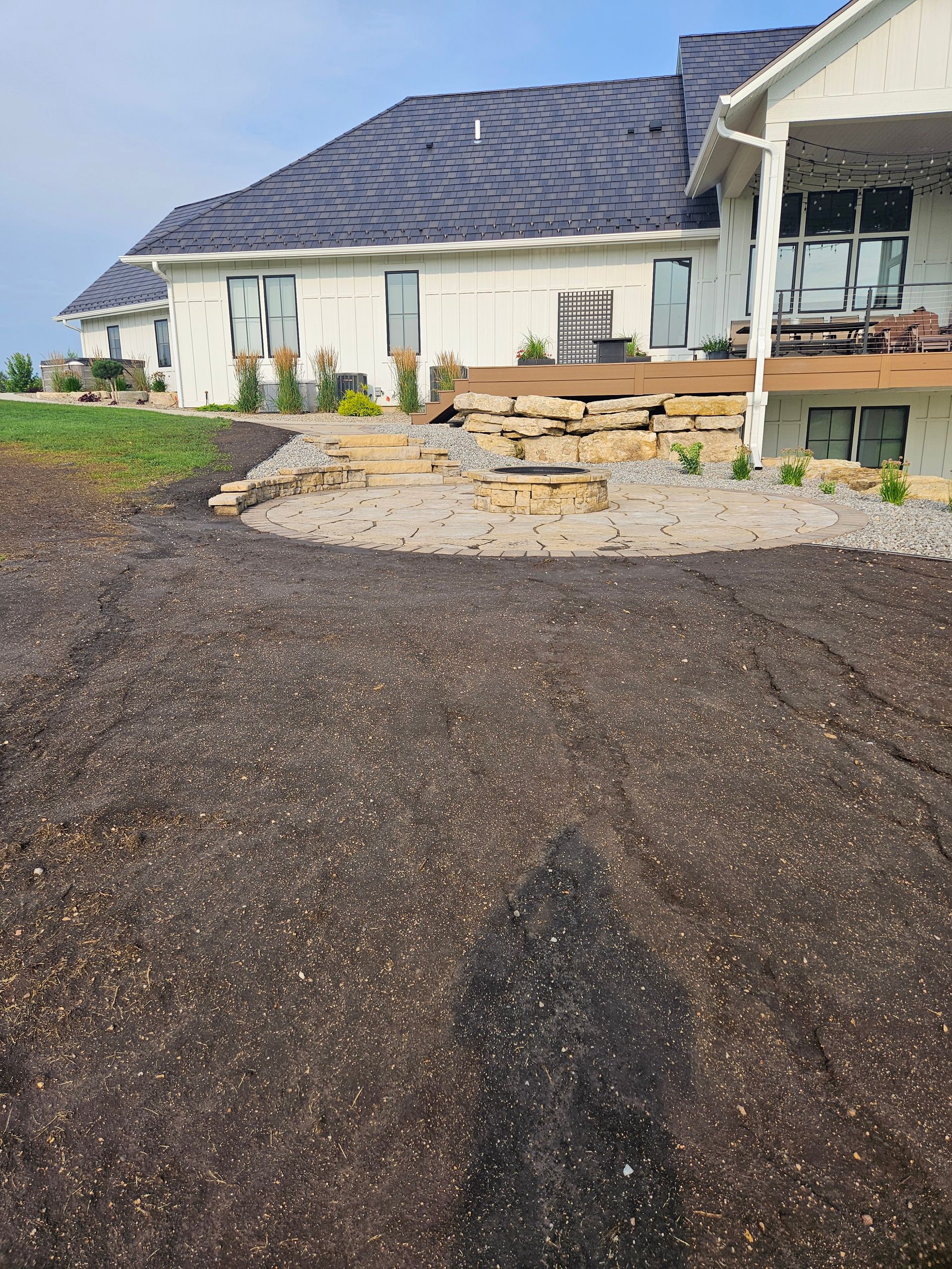 A freshly graded dirt yard leading to a stone fire pit area and a modern white house with a dark roof.