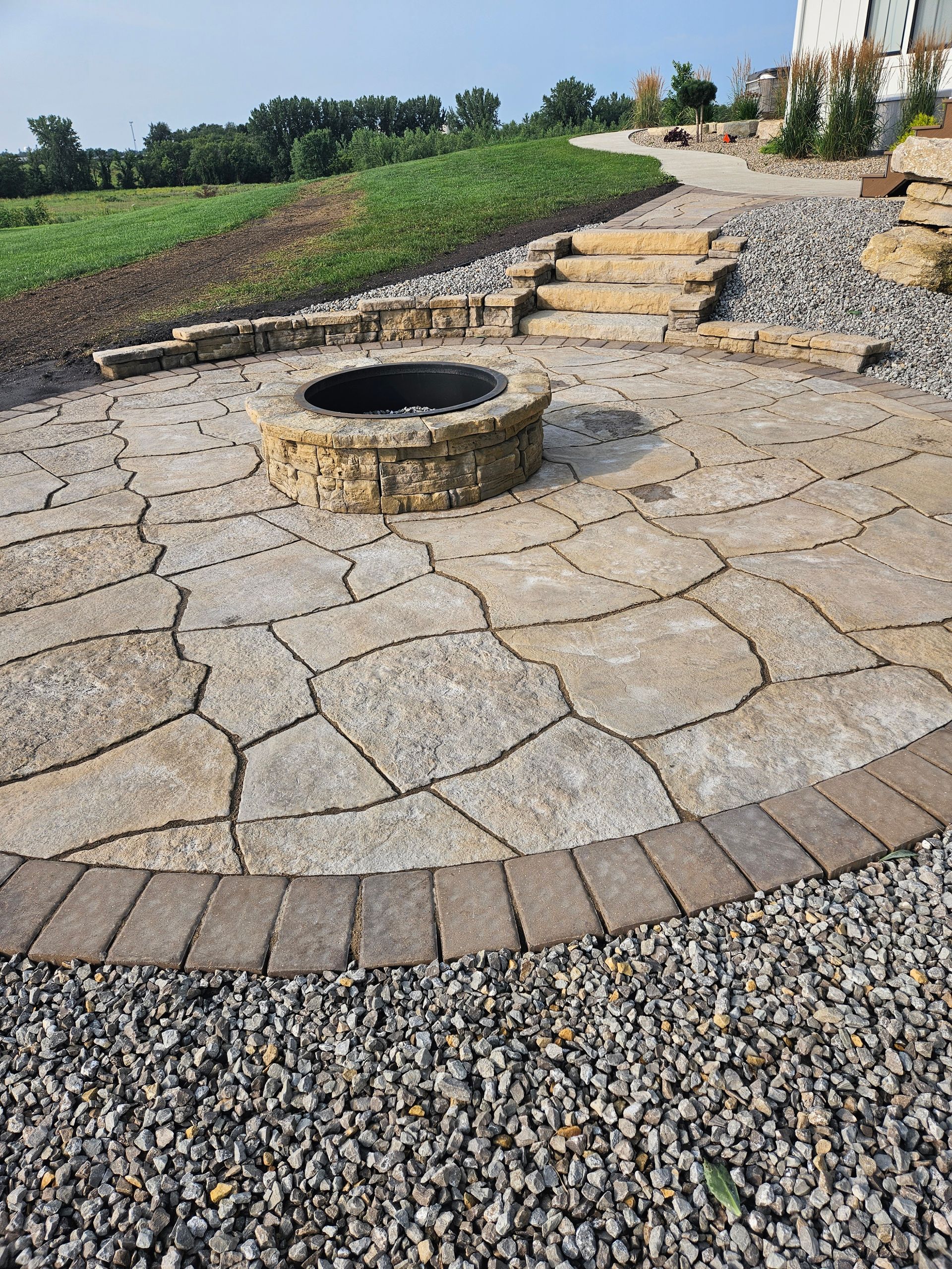 A circular stone fire pit patio with a stone border and steps leading to a gravel path in a landscaped yard.