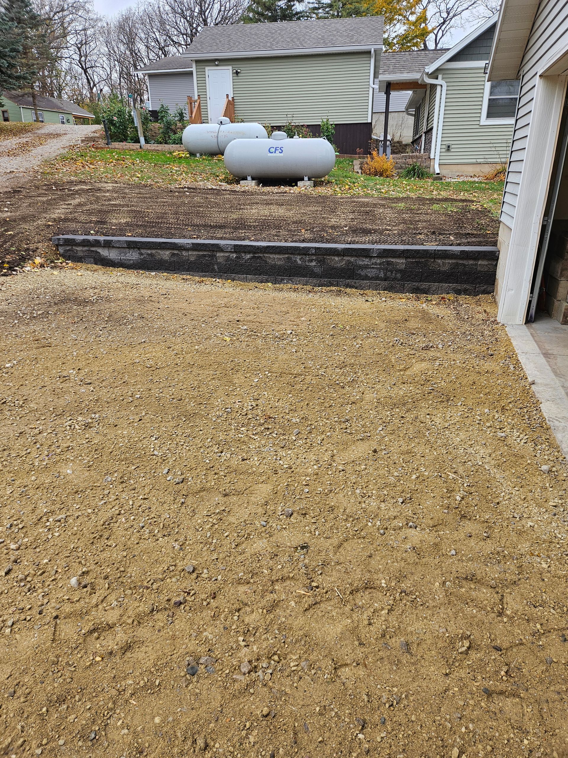 A yard with light-colored mulch in the foreground, a dark retaining wall, and two gray propane tanks near small houses.
