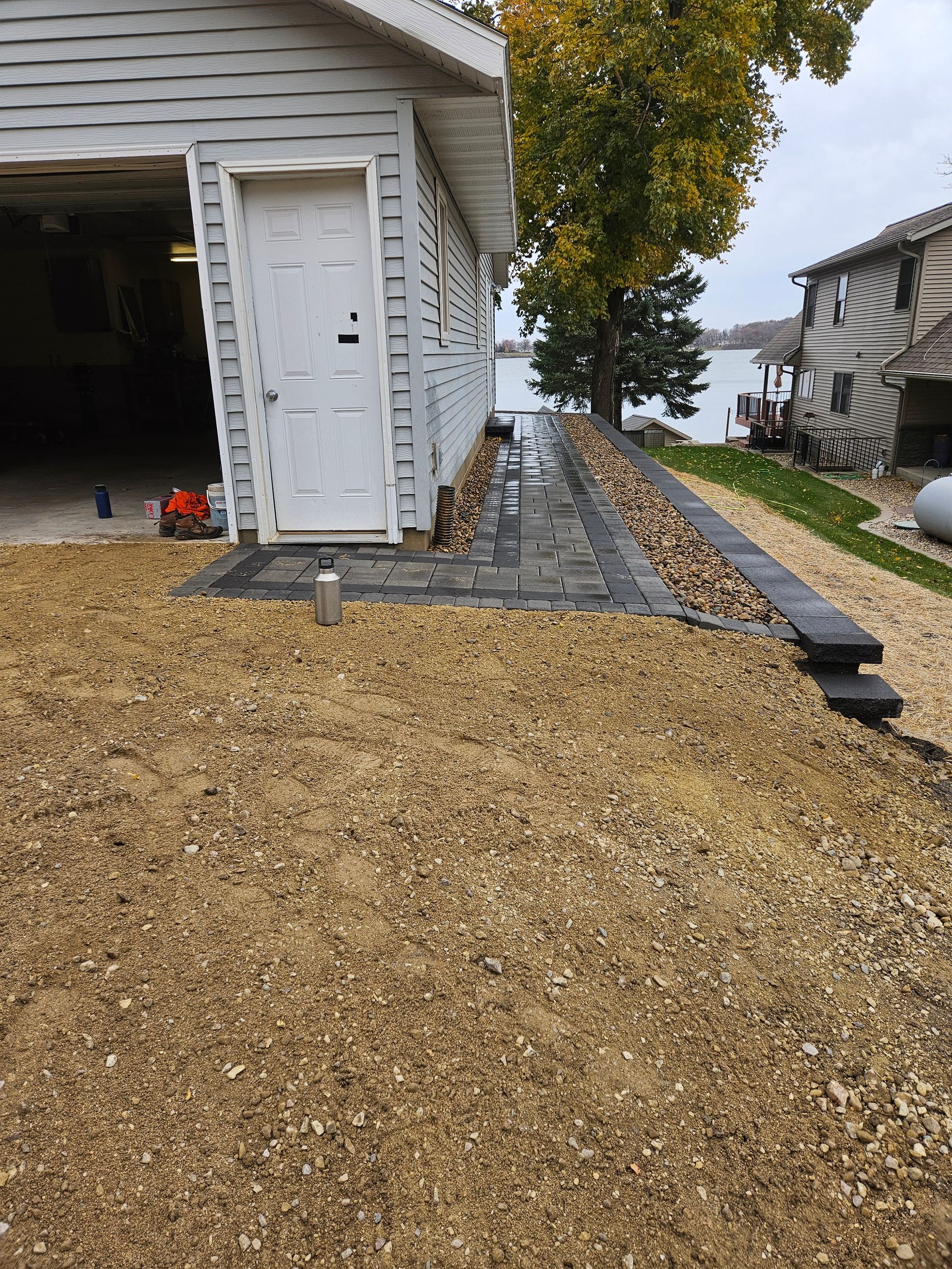 A white garage with a paver walkway leading to a lakefront view, surrounded by light-colored gravel landscaping.