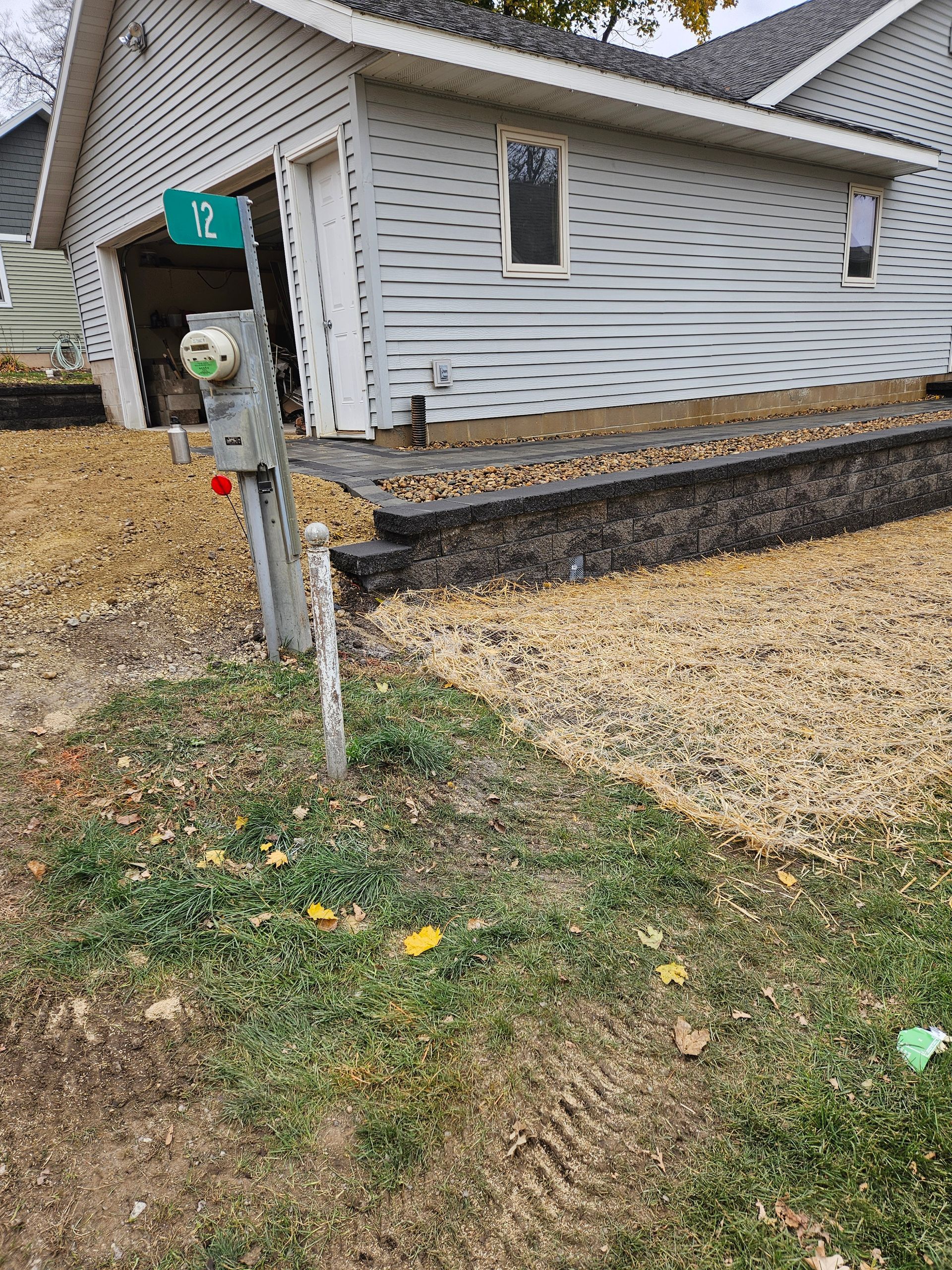 A grey-sided garage with a stone retaining wall, gravel yard, and a utility meter with a green sign labeled 