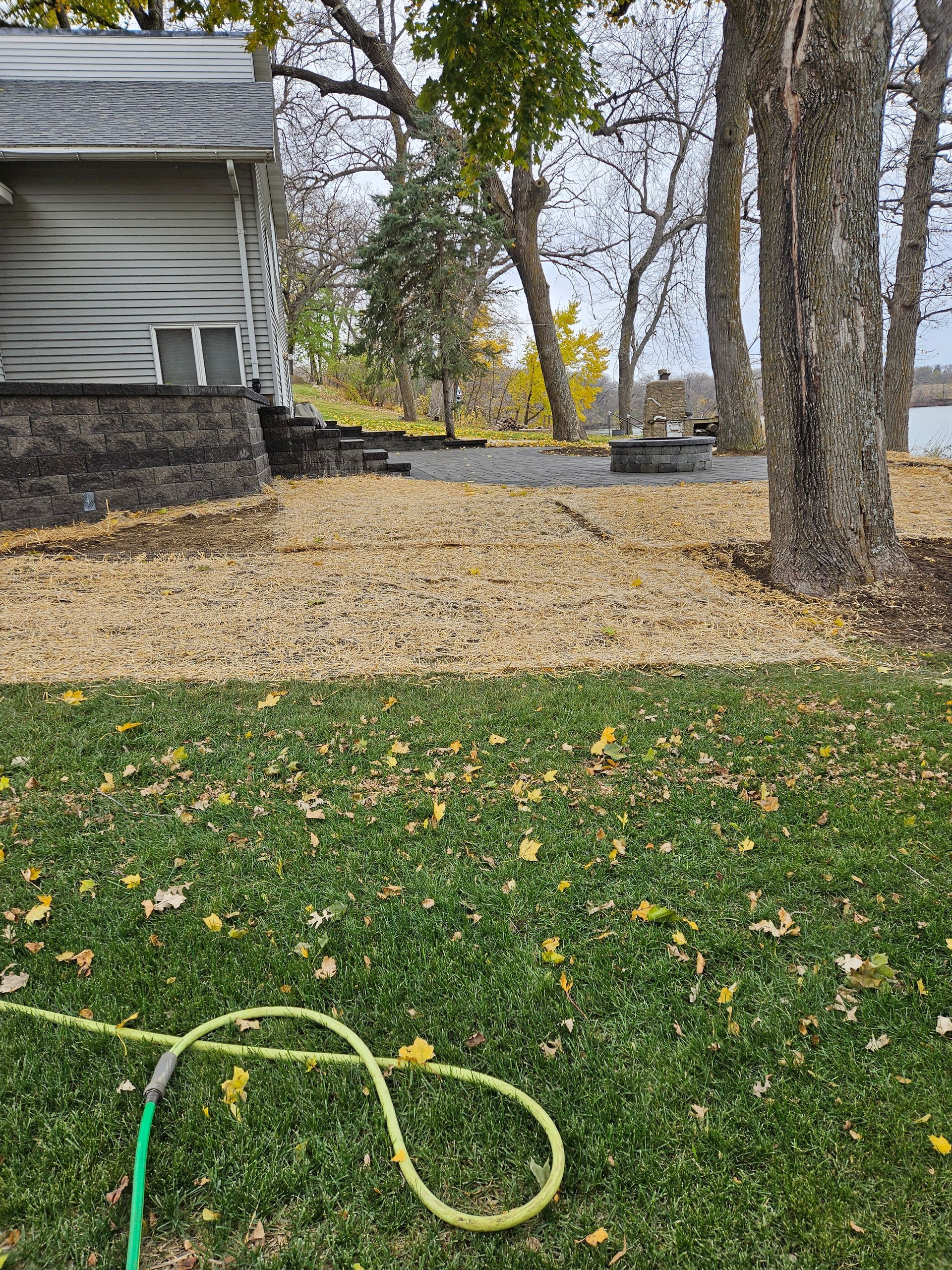 A yard covered in autumn leaves, showing a house corner, a stone patio with a fire pit, and a green garden hose in the grass.