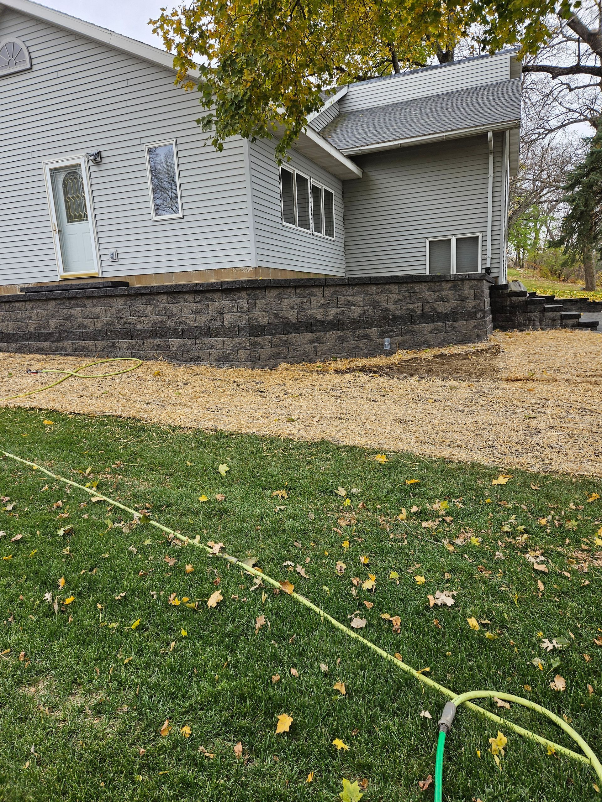 A gray house with white trim and a tiered stone retaining wall, set against a lawn with a garden hose in the foreground.