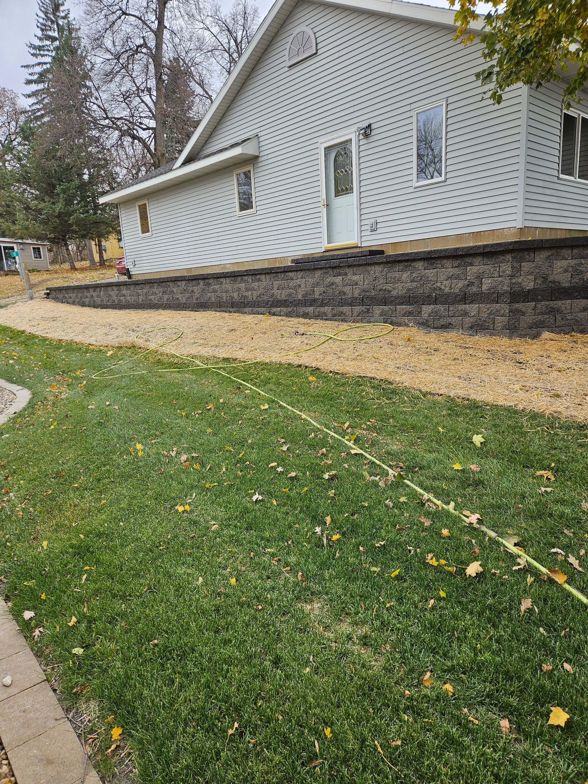A white house with gray siding sits behind a stone retaining wall and a gravel bed, overlooking a green, leaf-covered lawn.