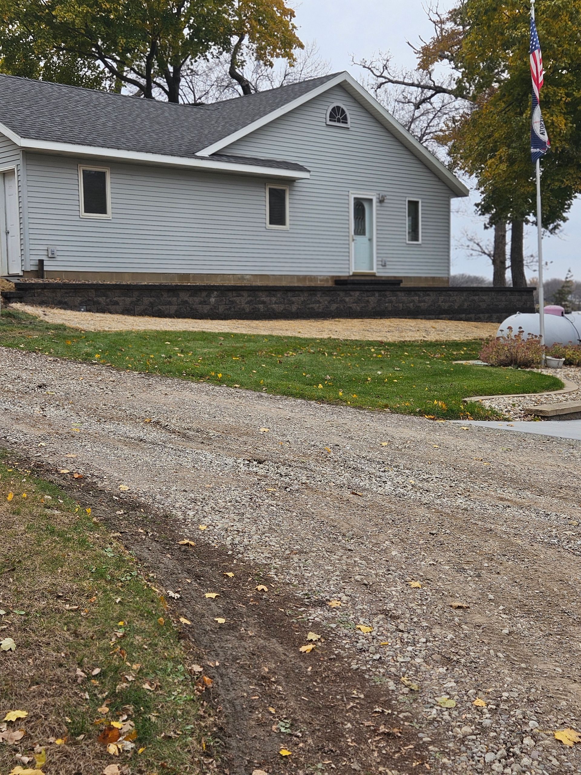 A light gray house with a stone retaining wall, set behind a gravel driveway and green lawn under an overcast sky.