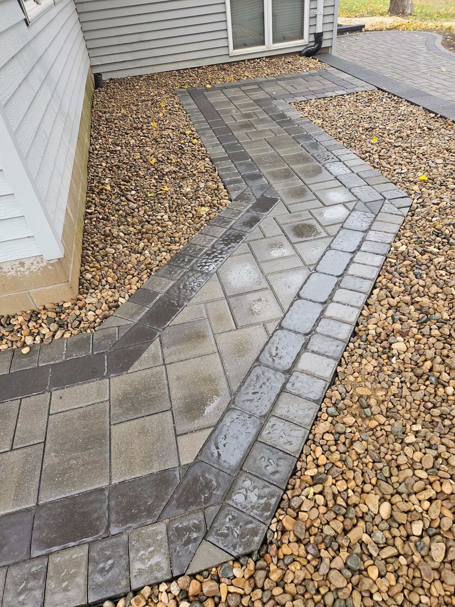 A grey paver walkway with a dark border cuts through a bed of tan landscape gravel beside a house with white siding.